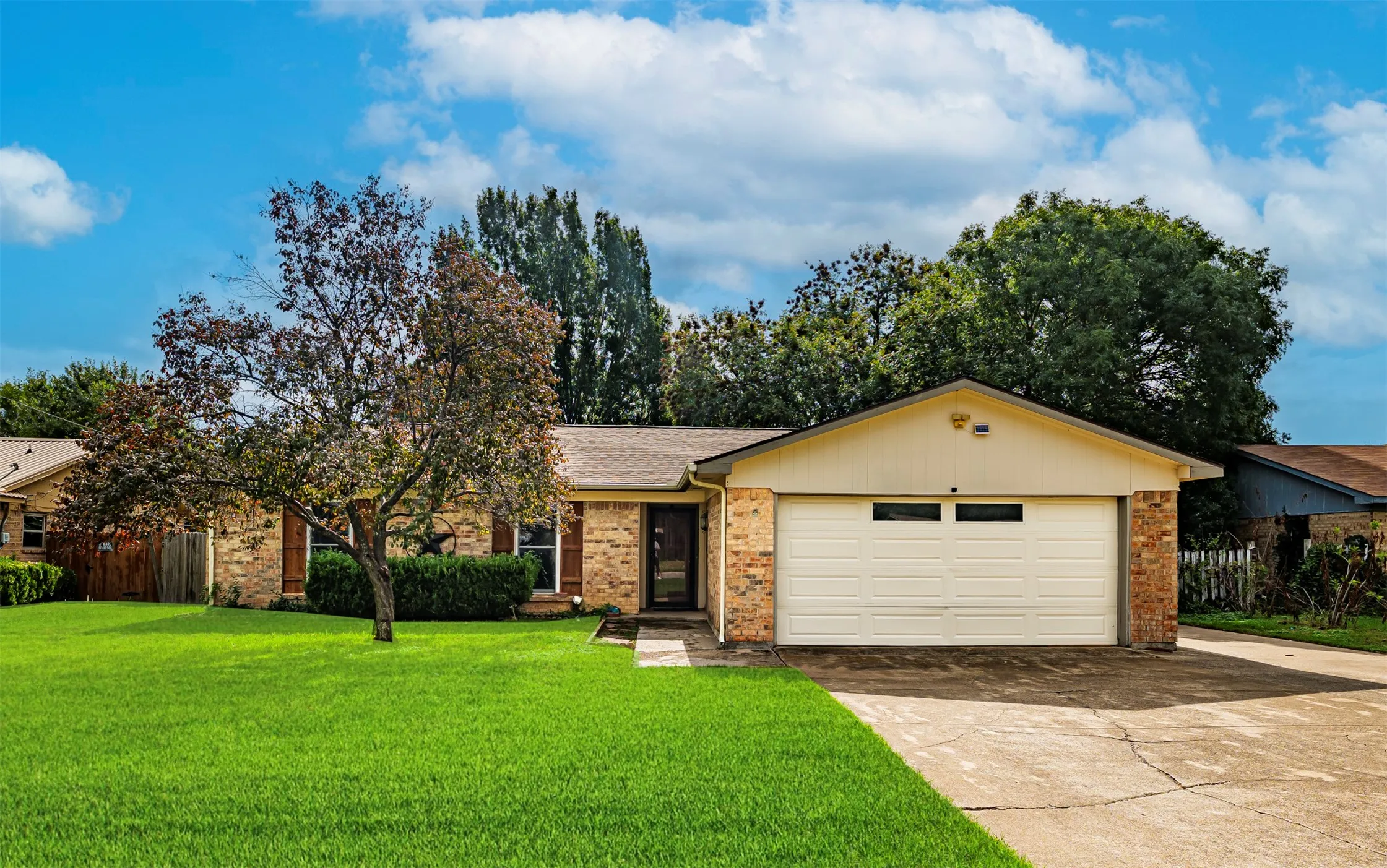 Single-story brick exterior featuring a front-facing attached two-car garage, manicured lawn, mature trees, and a concrete driveway