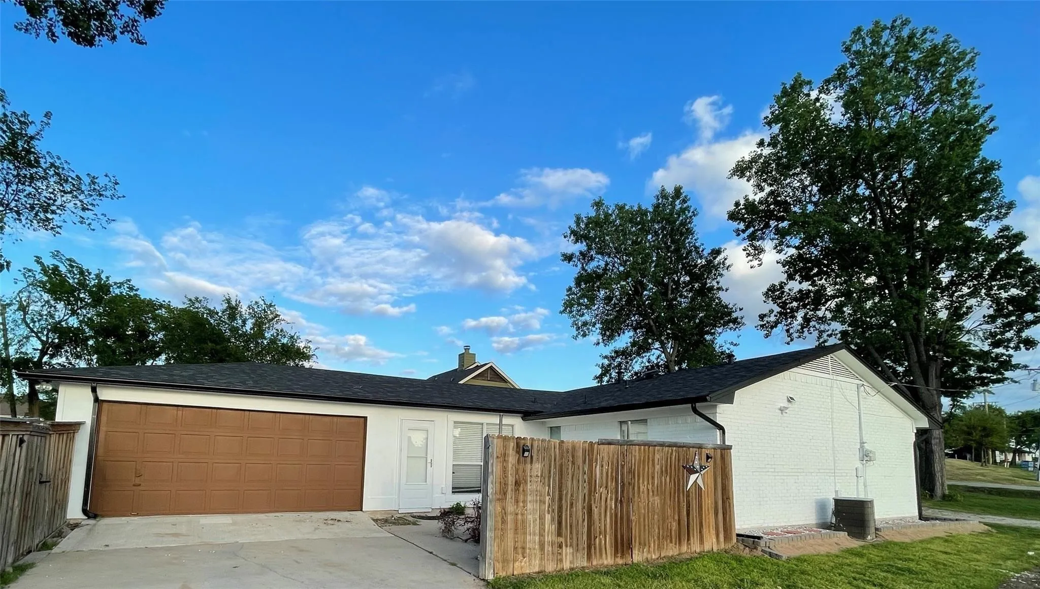 Exterior featuring white painted brick masonry, a dual-car garage with a paneled door, a dark composition shingle roof, and a wood privacy fence