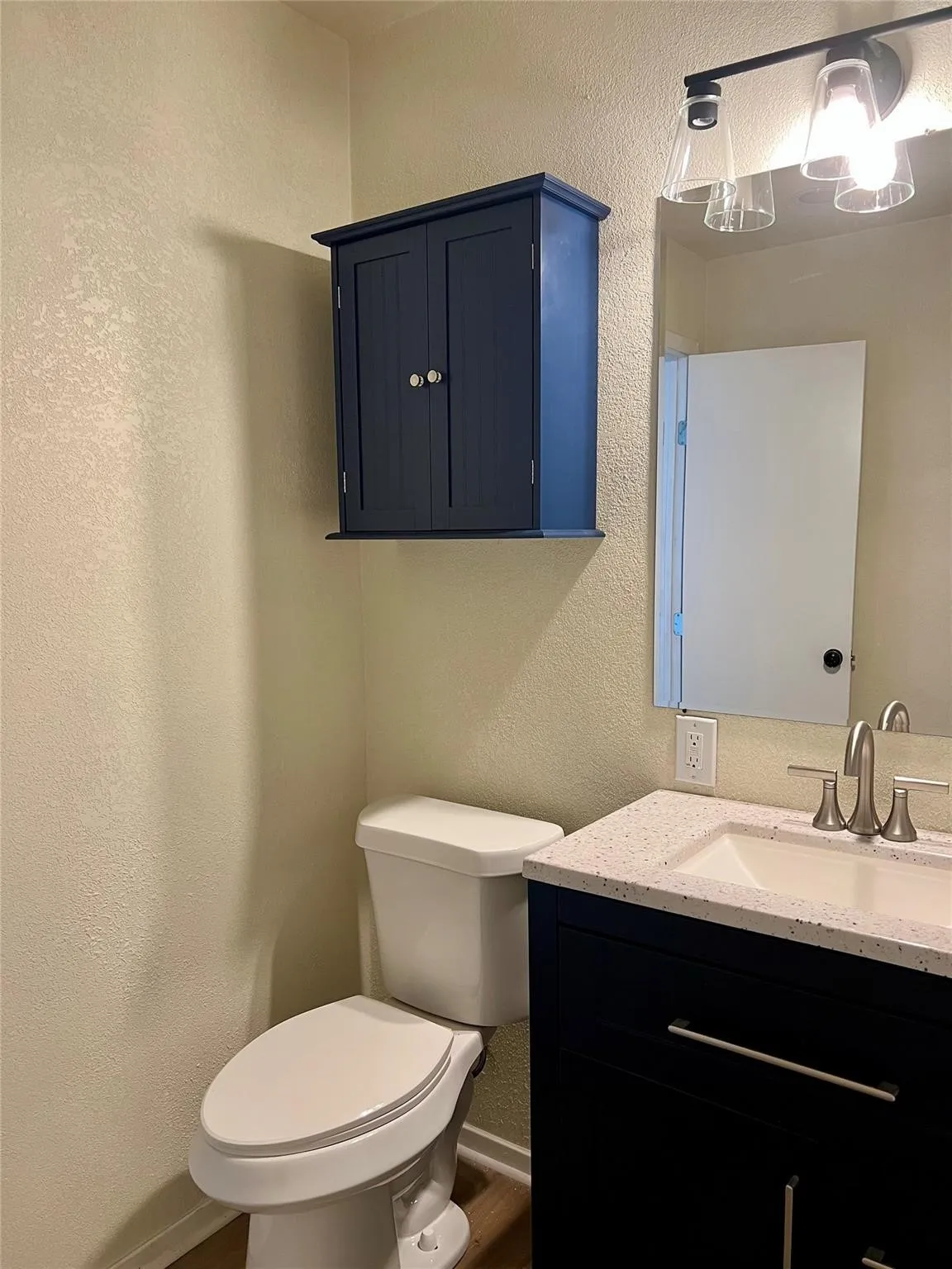 Navy blue vanity cabinet with a speckled countertop, integrated sink, brushed-nickel faucet, matching wall-mounted storage cabinet, and three-light overhead fixture