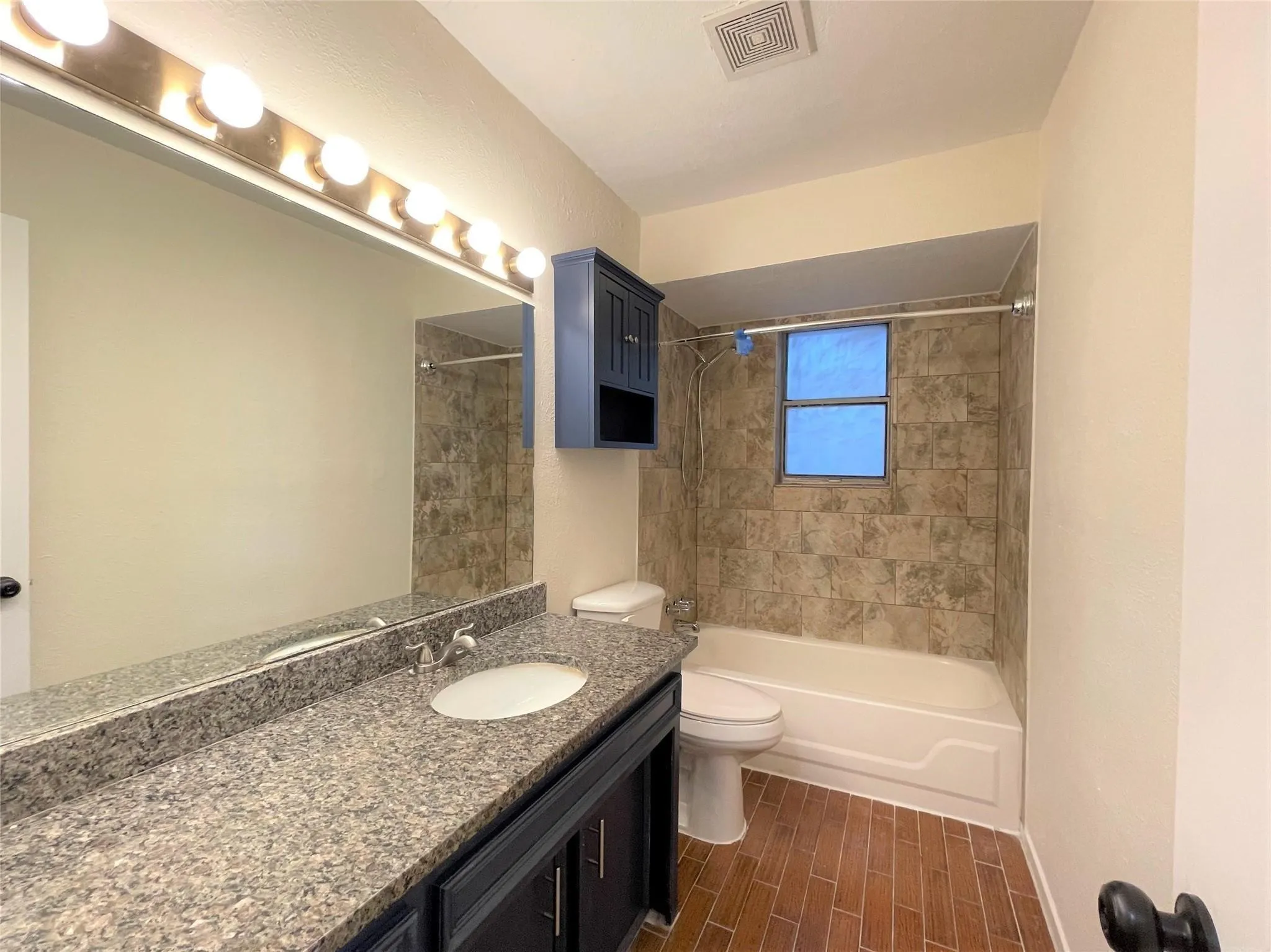 Bathroom featuring a vanity with speckled stone-finish countertop and oval undermount sink, dark cabinetry, wood-finish tile flooring, and tub surround with decorative wall tile
