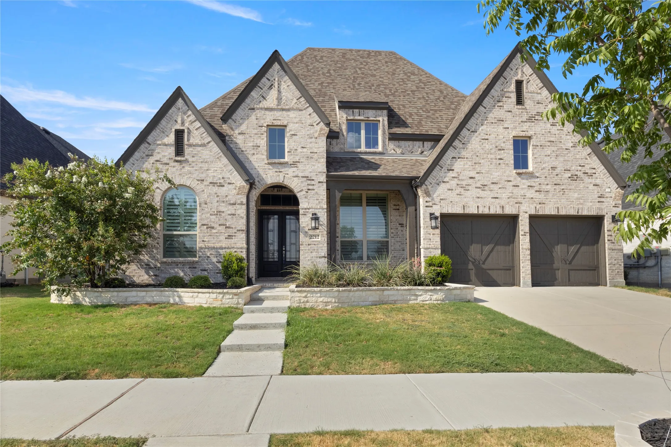 Traditional brick facade featuring multiple roof gables, arched entry, stone-accented landscape borders, and double-bay garage with decorative carriage-style doors