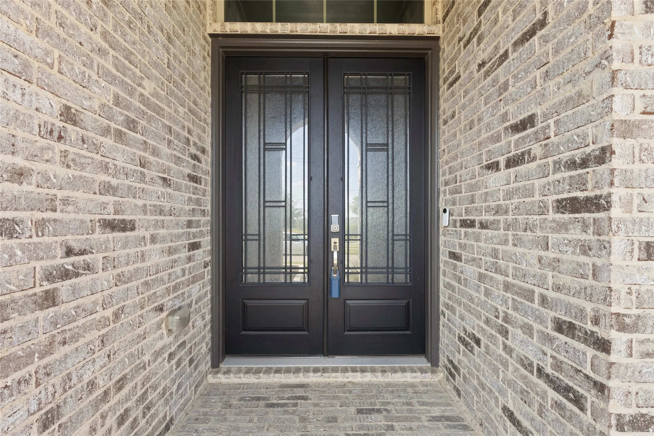 Double entry doors featuring dark-toned frames, geometric glass inserts, and a transom window, surrounded by neutral-toned brick masonry