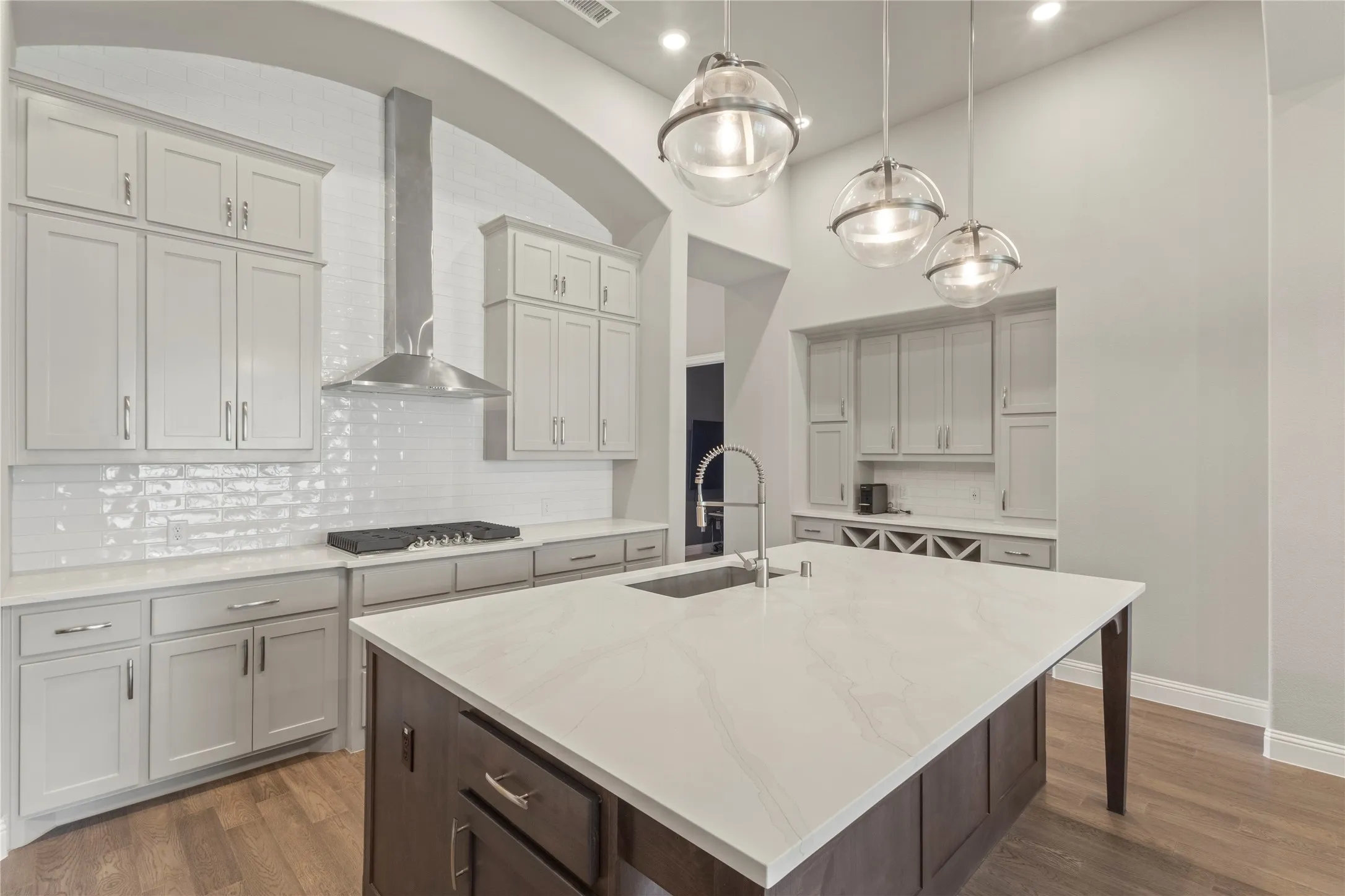 Kitchen featuring an expansive island with a light-toned countertop, custom cabinetry, stainless steel range hood, glass globe pendant lighting, and wood-finish flooring