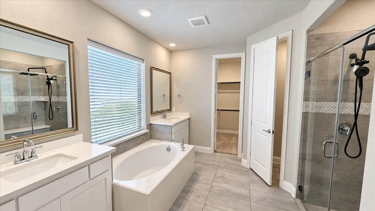 Bathroom featuring dual vanities with white cabinetry and solid-surface countertops, a deep soaking tub, a walk-in shower with tile surround, and a walk-in closet