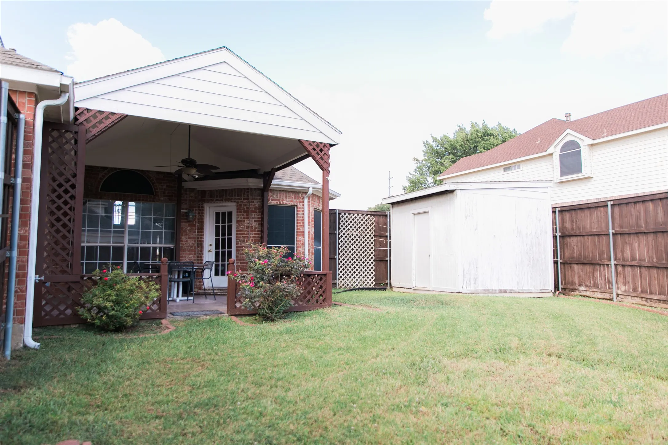 Covered patio featuring a peaked roof, attached ceiling fan, and decorative lattice accents, complemented by a fenced backyard and a standalone storage structure
