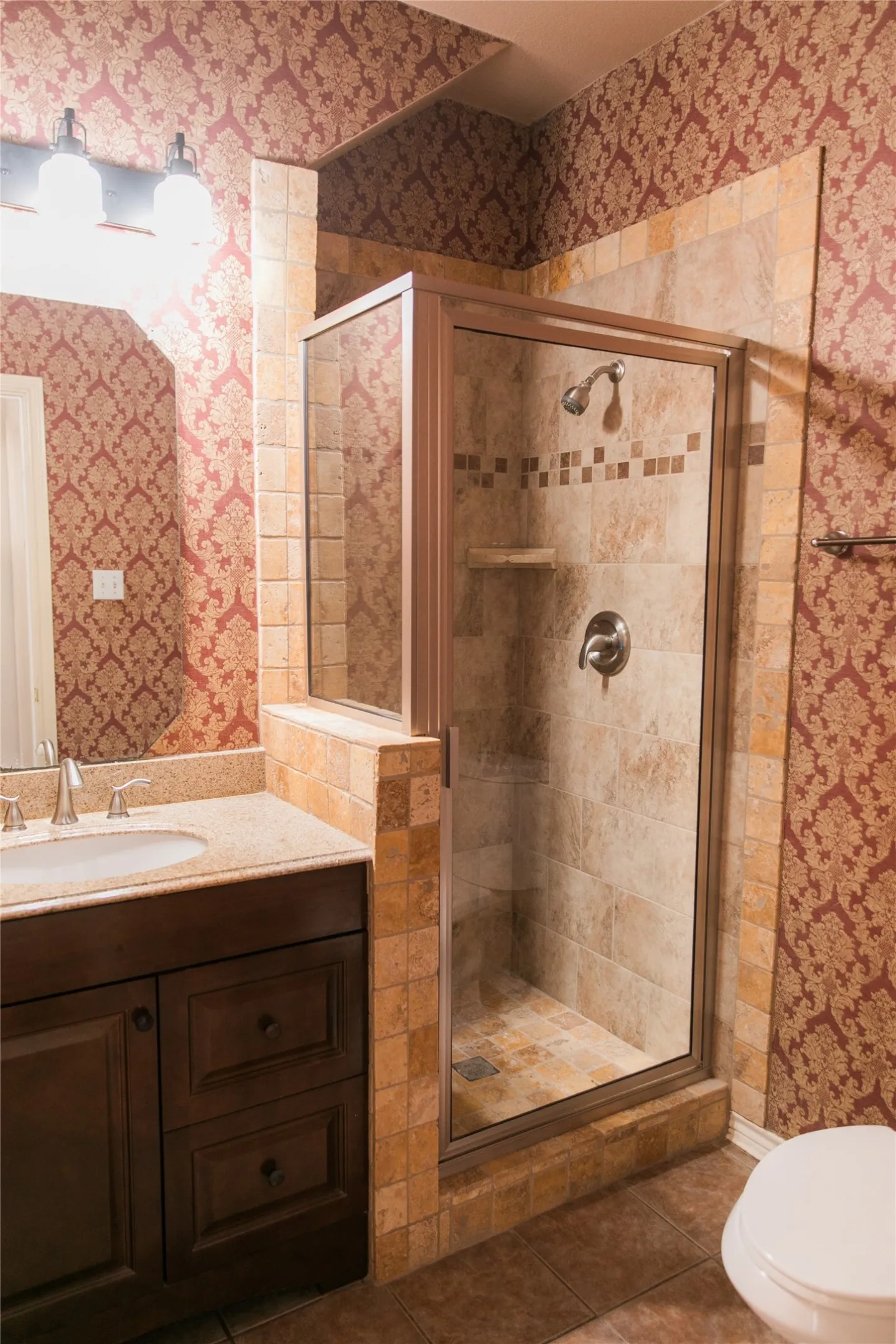 Dark wood-finish vanity with a stone countertop, integrated sink, shower enclosure with tiled surround, and decorative wall covering
