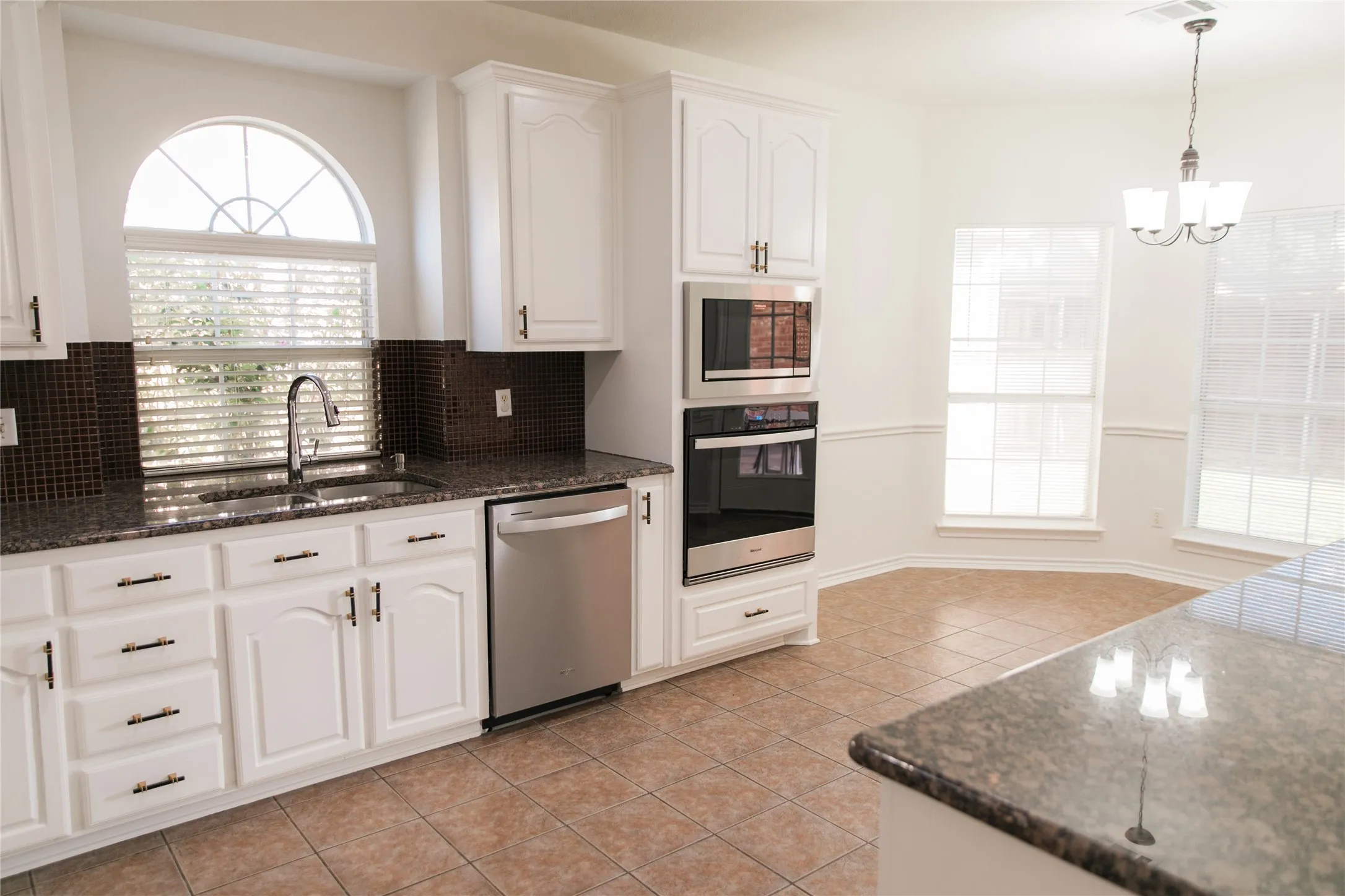 Kitchen featuring white shaker-style cabinetry, stone-surface countertops, a dark mosaic tile backsplash, stainless steel built-in double ovens, and ceramic tile flooring