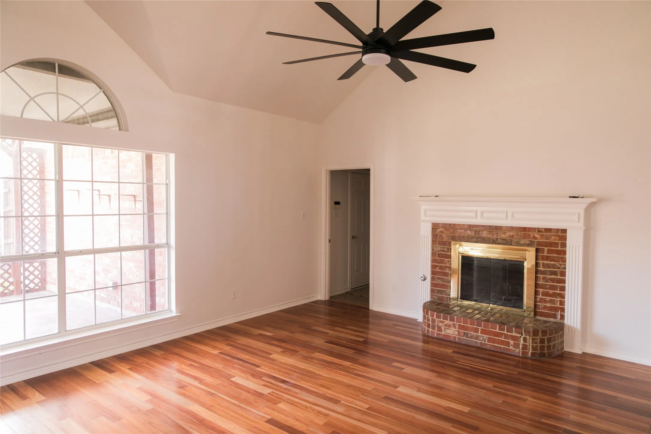 Vaulted ceiling with a matte black multi-blade ceiling fan, wood-finish flooring, brick fireplace with a white mantel, and an expansive window featuring an arched transom