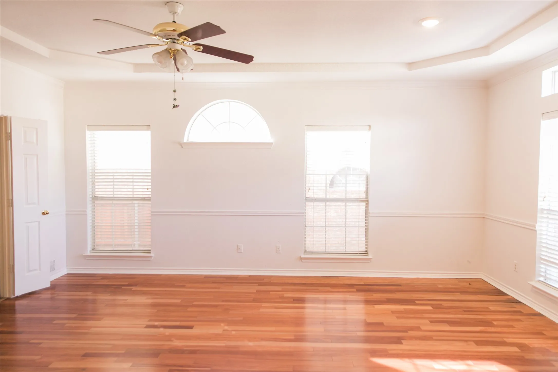 Wood-finish flooring, tray ceiling, ceiling fan with integrated lighting, arched window, and chair rail molding
