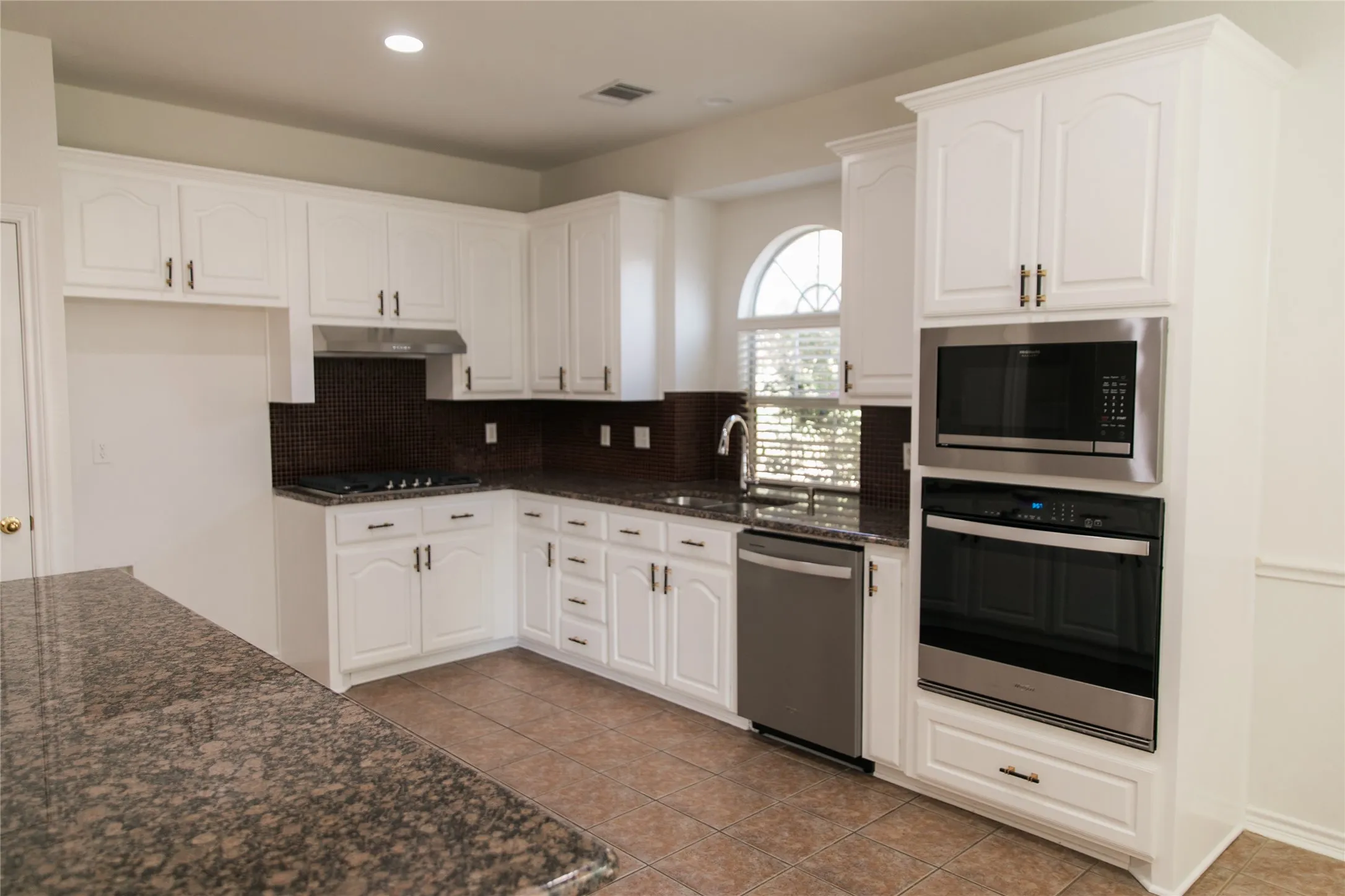 Kitchen featuring white cabinetry, stone-finish countertops, dark tile backsplash, stainless steel built-in double oven and microwave, and an arched window