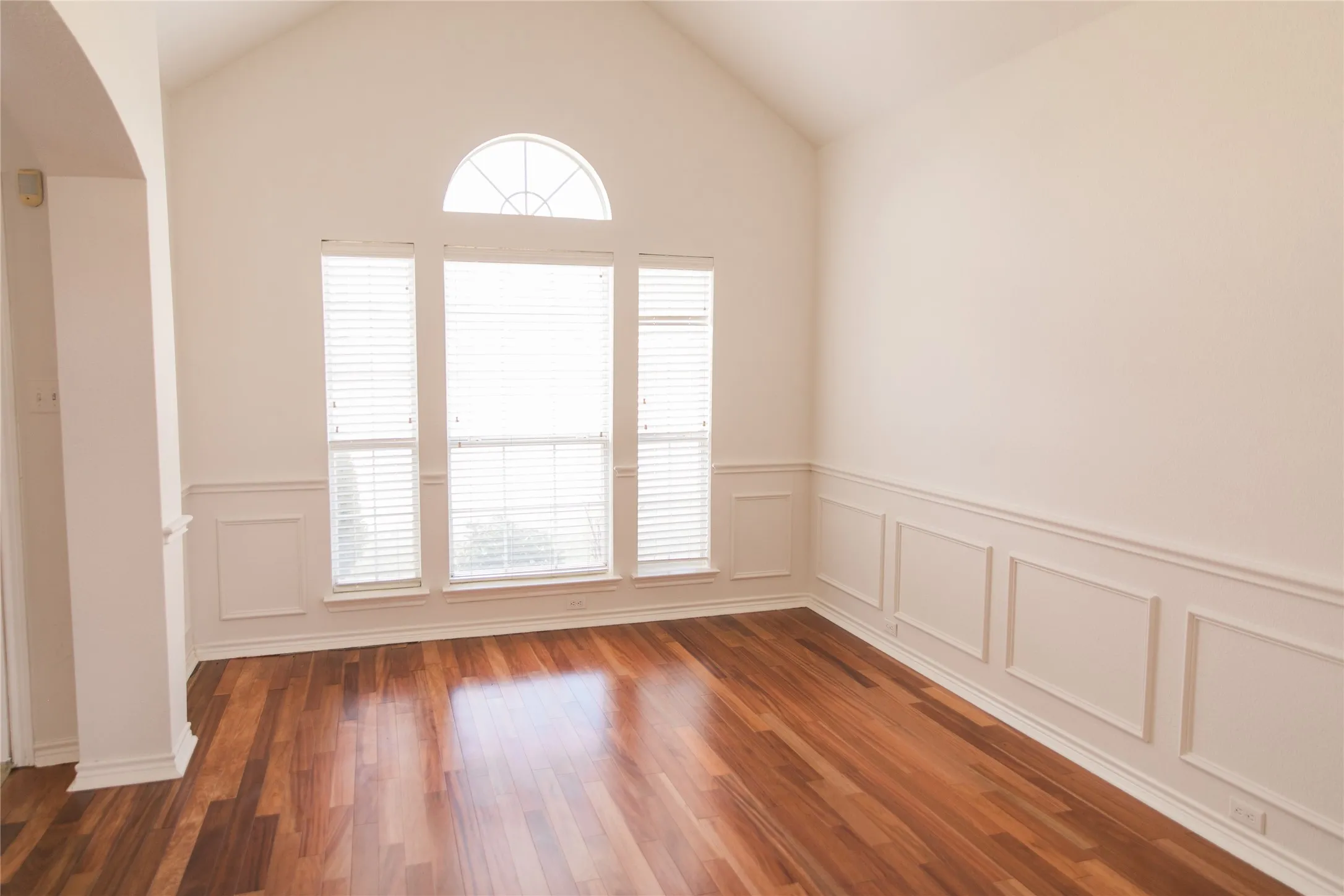 Vaulted ceiling, wood-finish flooring, and white wainscoting complemented by triple-panel windows featuring a prominent arched transom