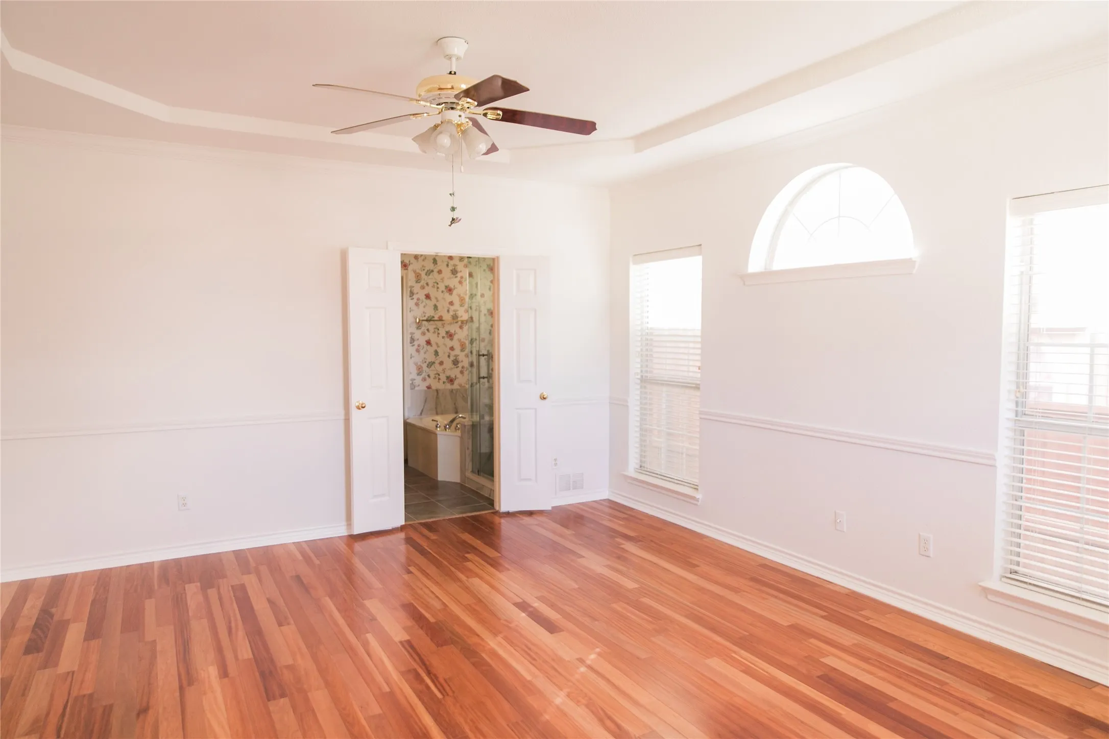 Room featuring wood-finish flooring, tray ceiling, ceiling fan with integrated lighting, dual-pane windows with a fixed arched transom, and chair rail molding
