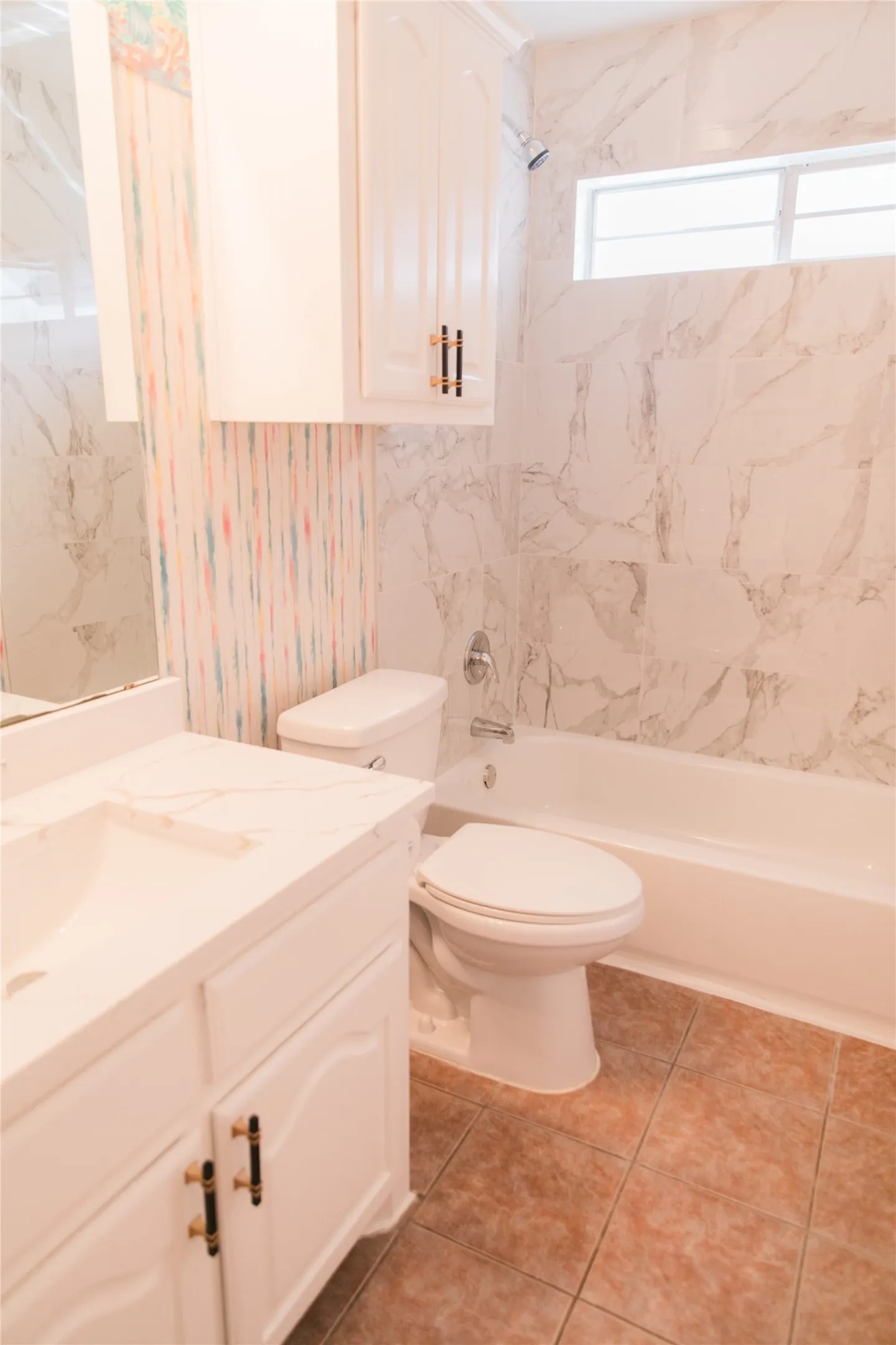Bathroom featuring a vanity with stone-finish countertop, white cabinetry with contrast hardware, full-height wall tiling, integrated bathtub, and upper transom window