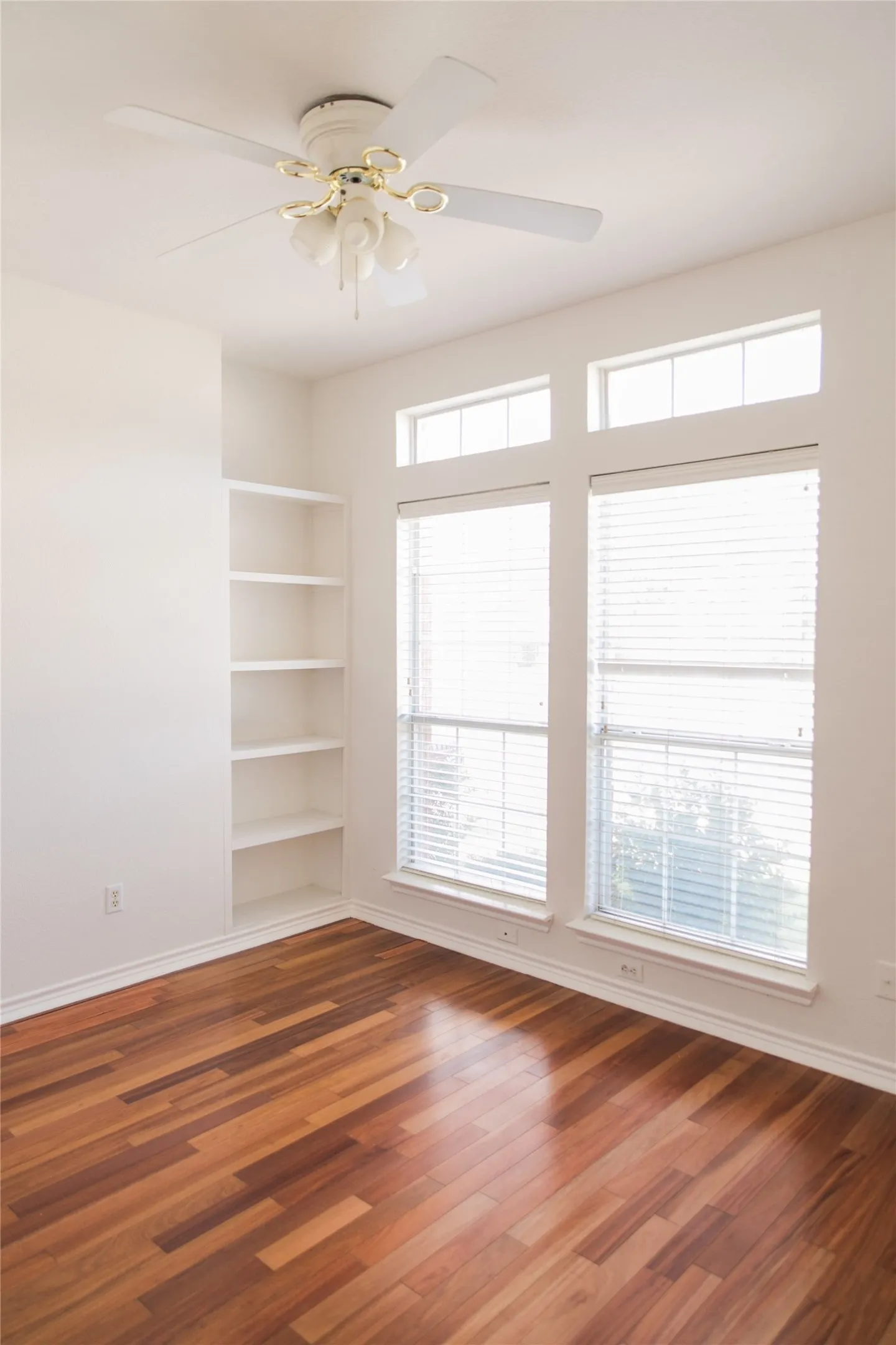 Wood-finish flooring, built-in shelving, dual windows with transom panes, and a ceiling fan with integrated lighting