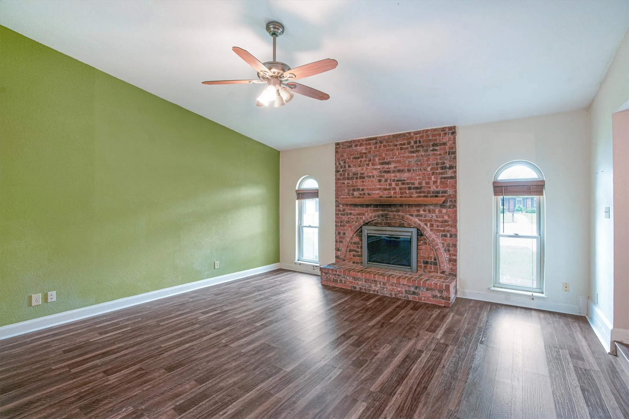 Vaulted ceiling featuring a ceiling fan, exposed brick fireplace with an arched hearth, wooden mantel, and luxury vinyl plank flooring with arched windows