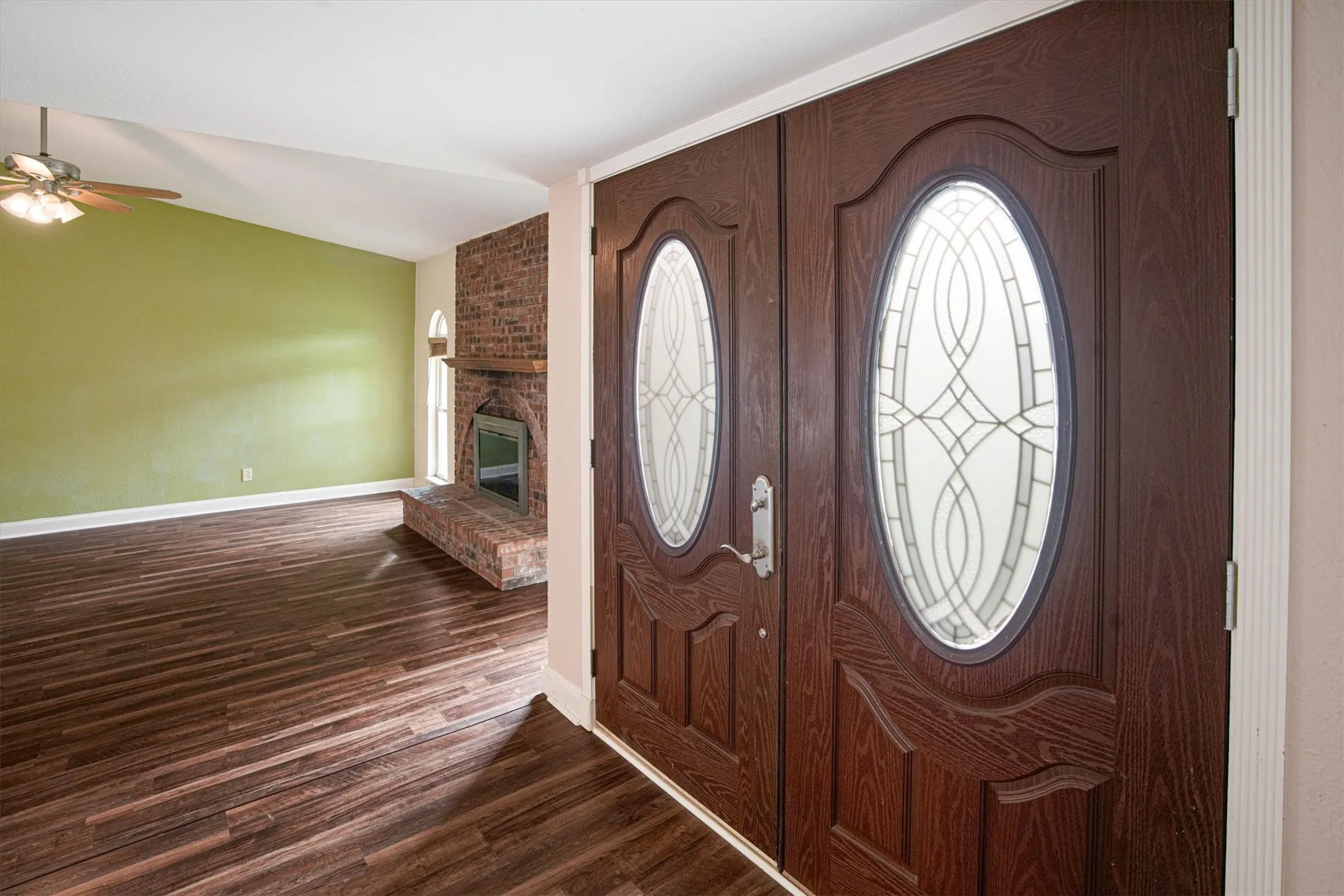Double entry doors featuring decorative oval glass inserts, luxury vinyl plank flooring, vaulted ceilings, and a floor-to-ceiling brick fireplace with a wood mantel