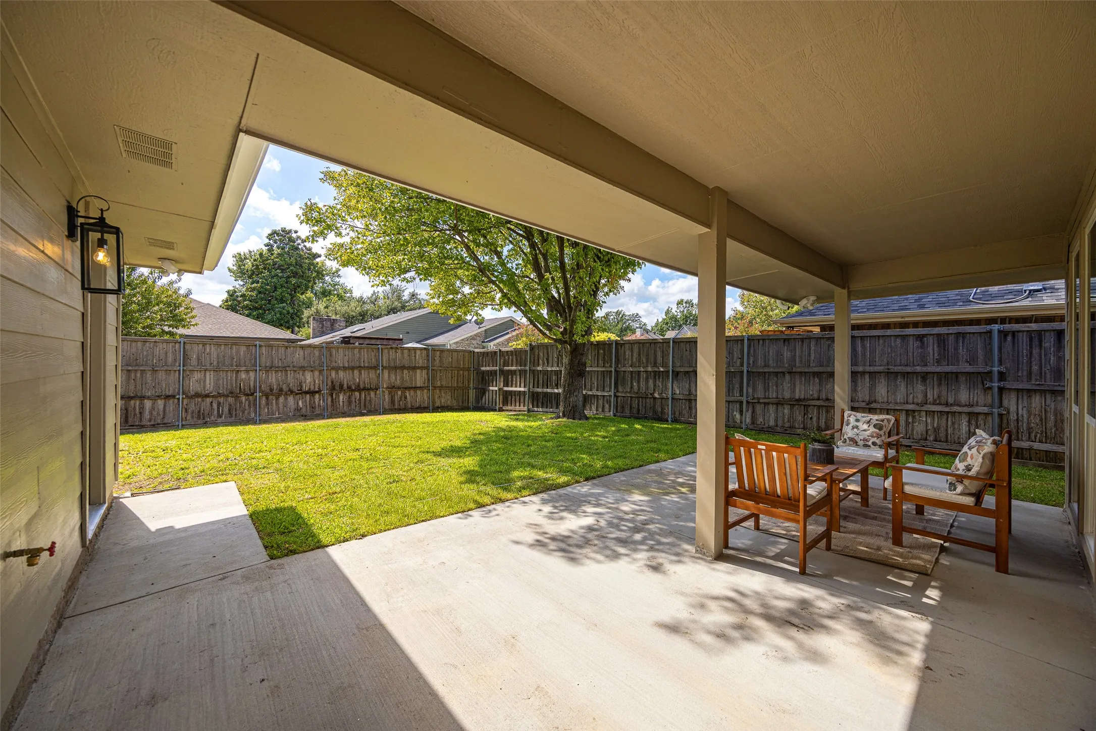Covered patio with generous space for outdoor seating and dining, overlooking the fenced backyard and mature shade tree.
