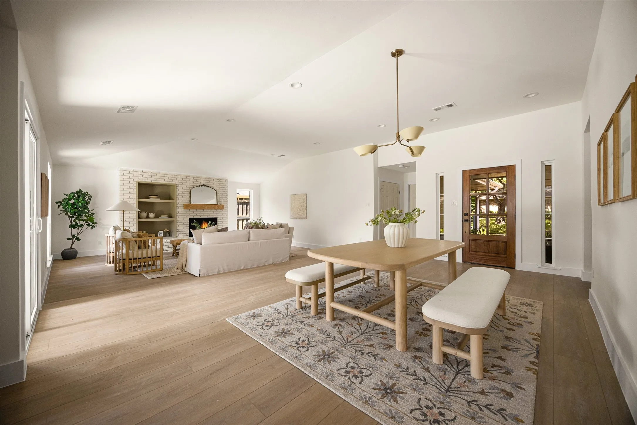 Open living and dining areas with vaulted ceilings, wood-like flooring, modern lighting, and a brick fireplace with built-in shelving.
