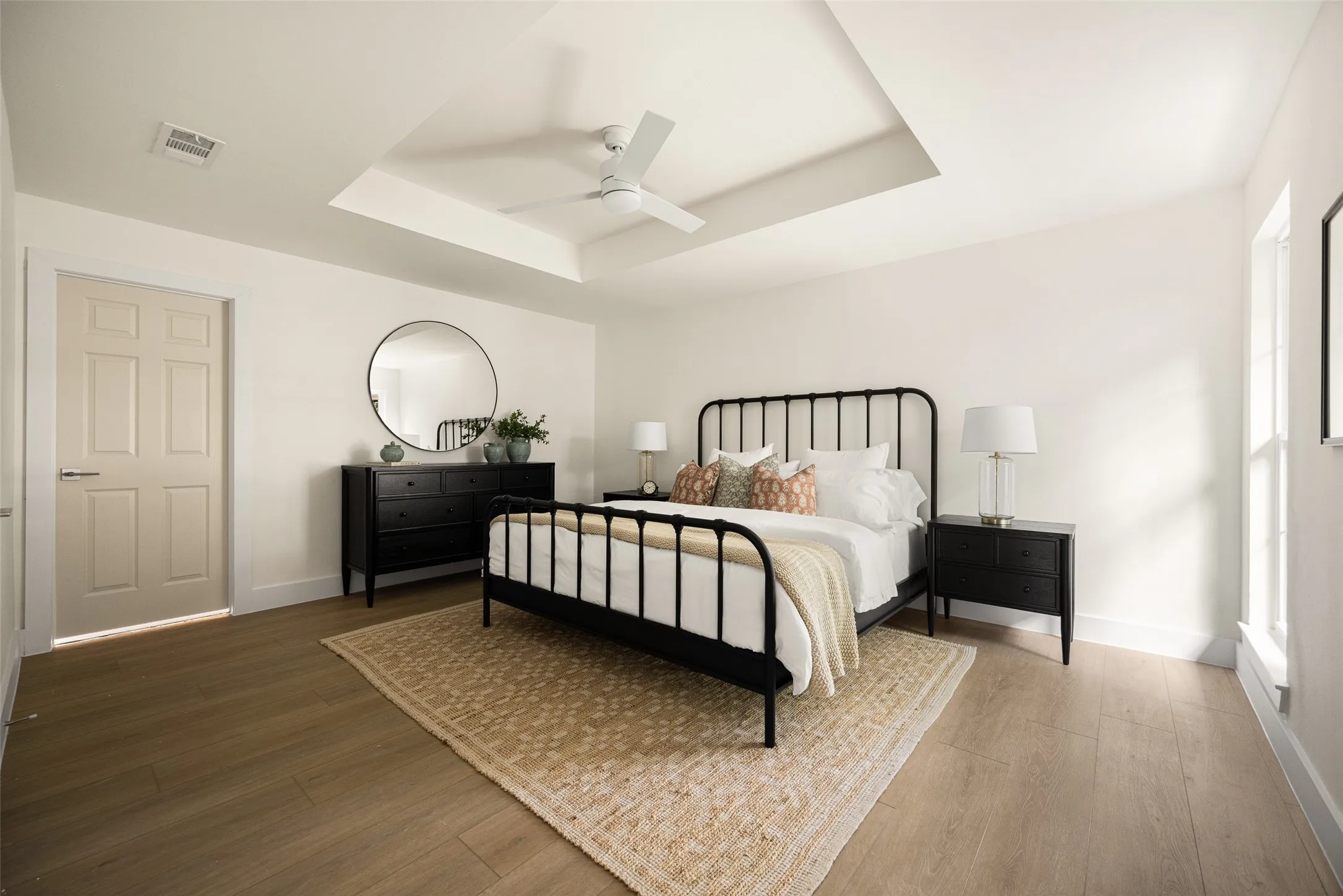 Beautifully updated primary bedroom featuring a tray ceiling, modern ceiling fan, warm wood-like flooring, and abundant natural light that creates a bright, inviting atmosphere