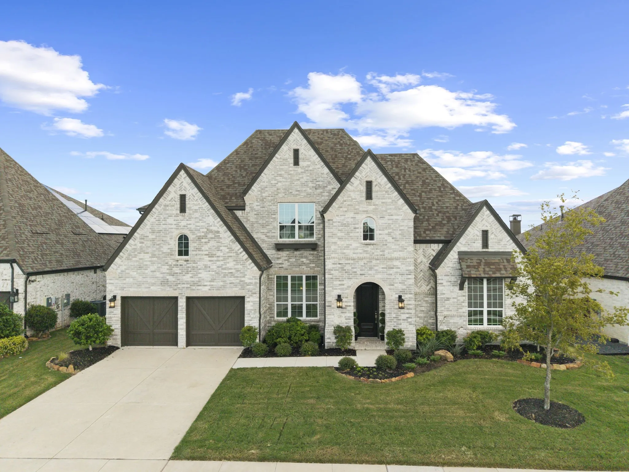 Stone and brick facade featuring multi-gabled rooflines, arched entryway, dark wood-finish garage doors, and manicured front landscaping