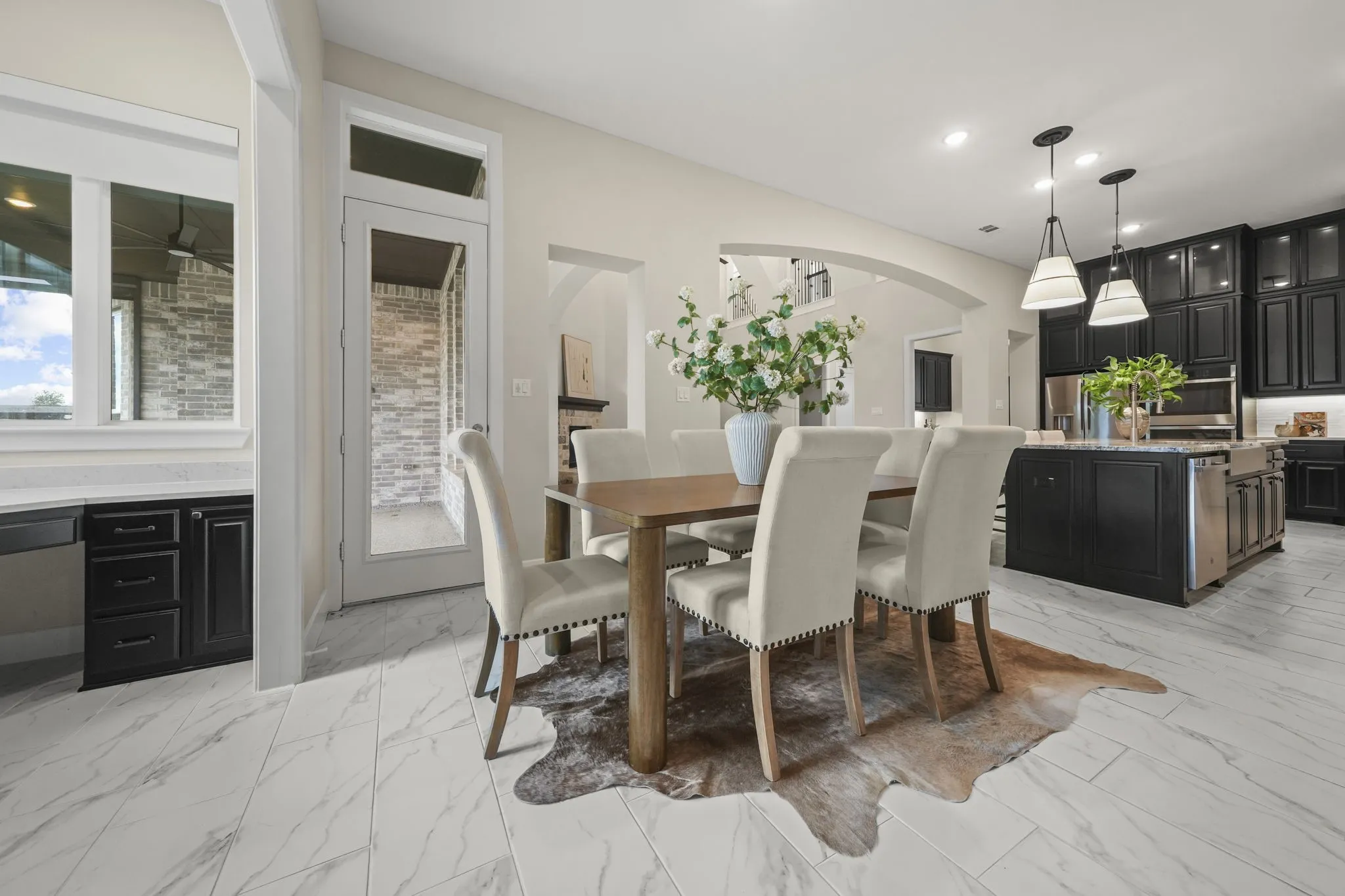 Open-concept kitchen and dining area featuring stone-look tile flooring, shaker-style cabinetry, a central island with an integrated sink, dual pendant lighting, and arched architectural details