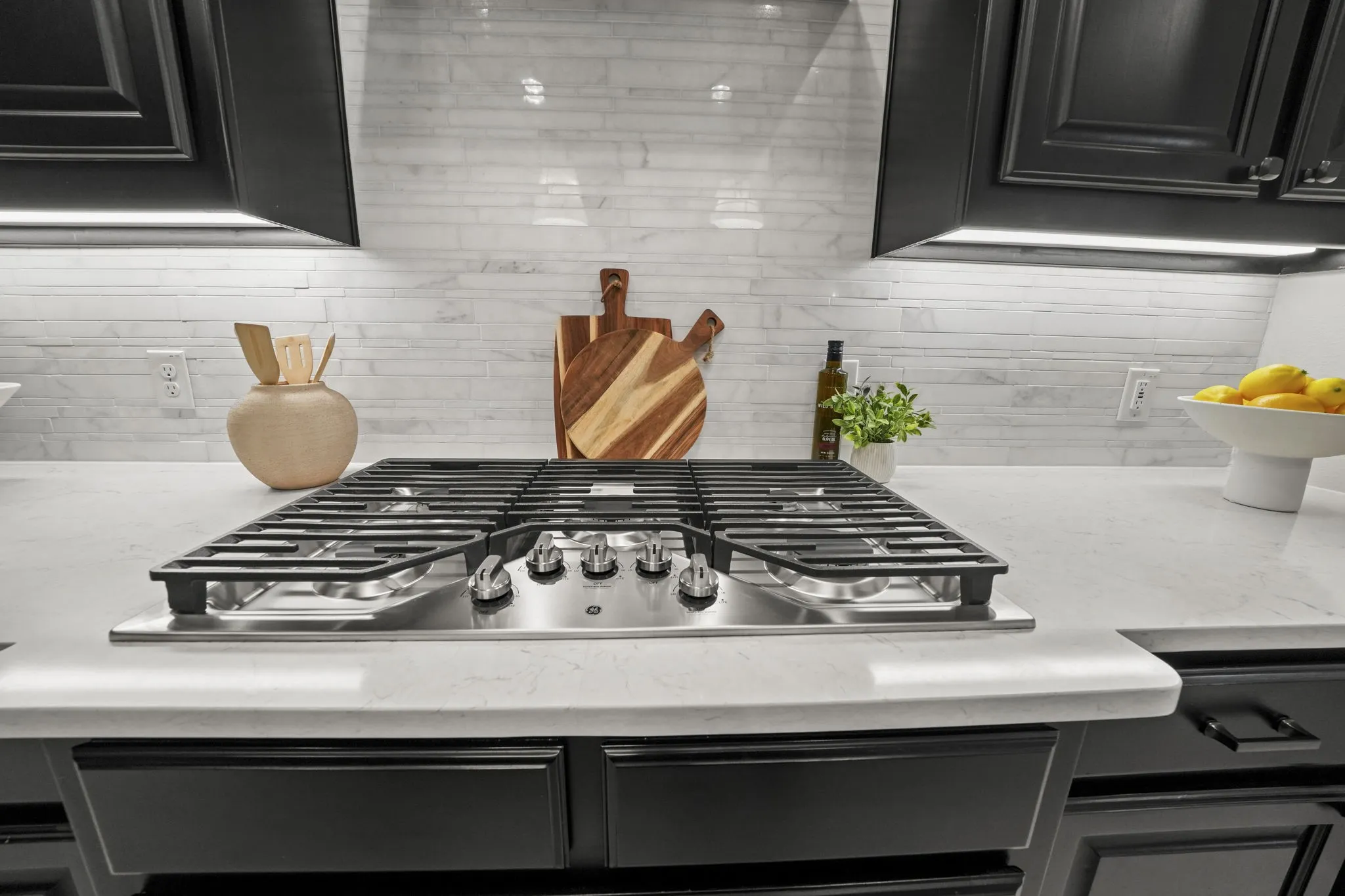 Stainless steel gas cooktop with heavy-duty grates, light-toned stone-finish countertops, linear tile backsplash, and dark shaker-style cabinetry with under-cabinet lighting