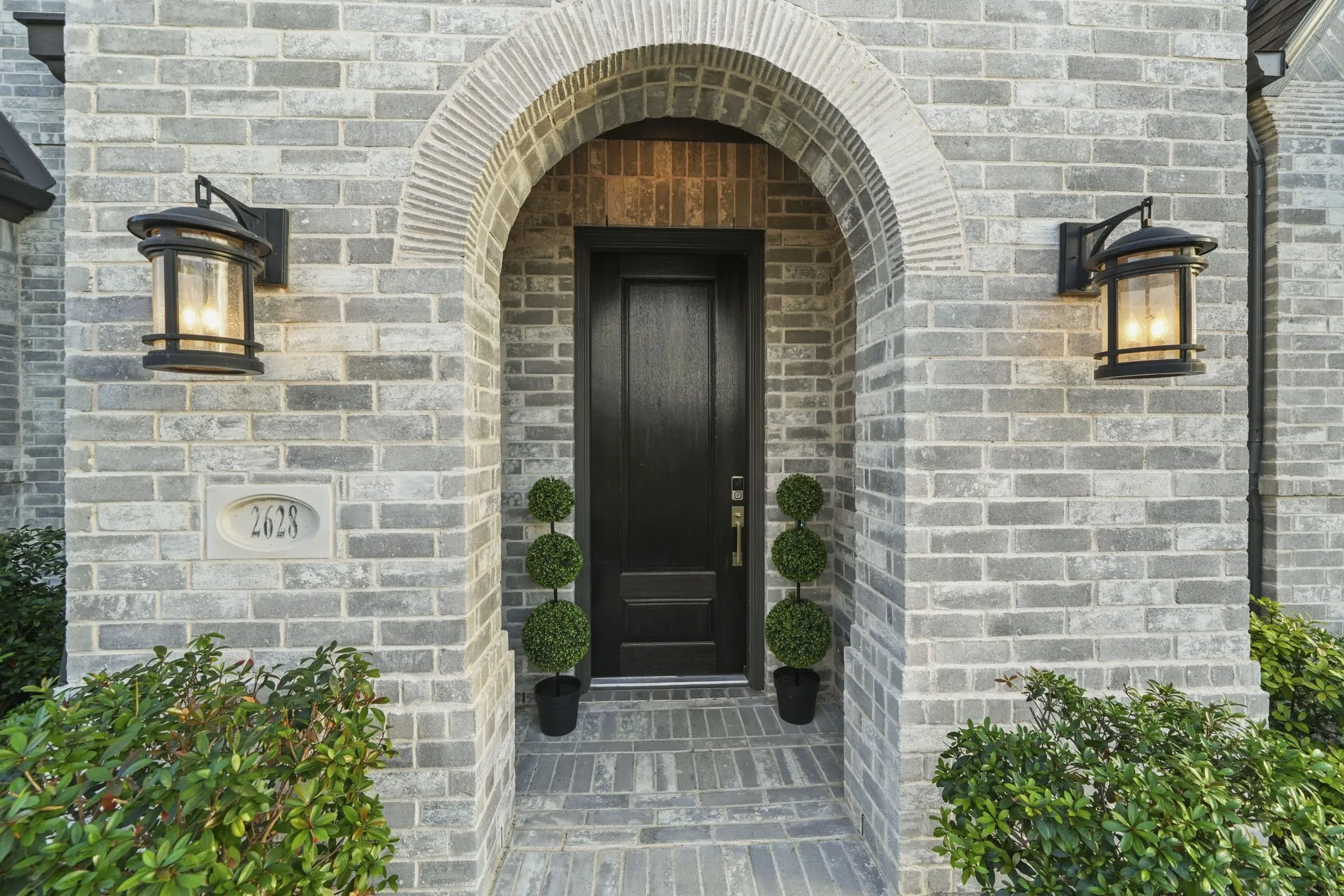 Entryway featuring a grey brick facade, arched stone surround, solid dark-finish entry door, and two wall-mounted metal lanterns