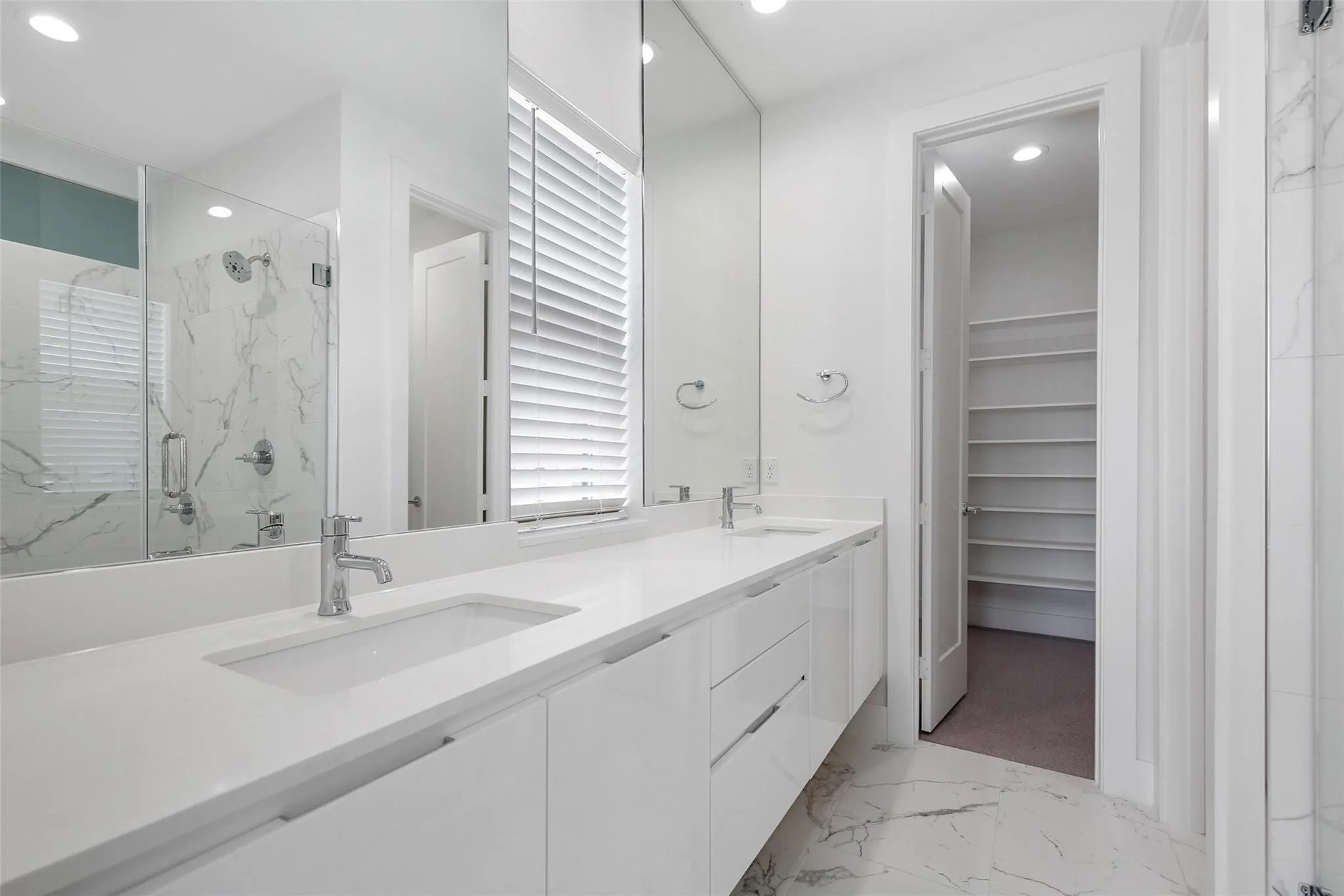 Floating white vanity with dual rectangular sinks, quartz countertops, floor-to-ceiling mirrors, large-format stone-finish tile flooring, and a walk-in storage area with custom shelving
