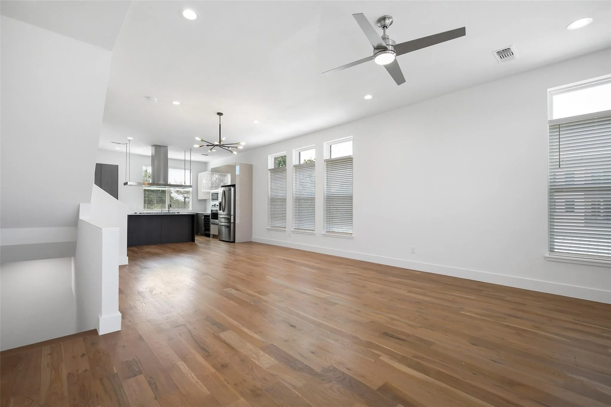 Open-concept interior featuring wood-finish flooring, recessed lighting, a modern ceiling fan, multiple windows with blinds, and a kitchen equipped with a stainless steel range hood