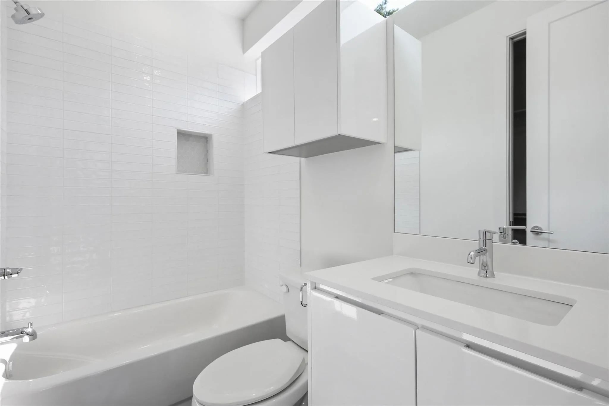 Bathroom featuring a white gloss vanity with integrated sink, stone-finish countertop, white tile tub surround with built-in recessed niche, and wall-mounted storage cabinetry