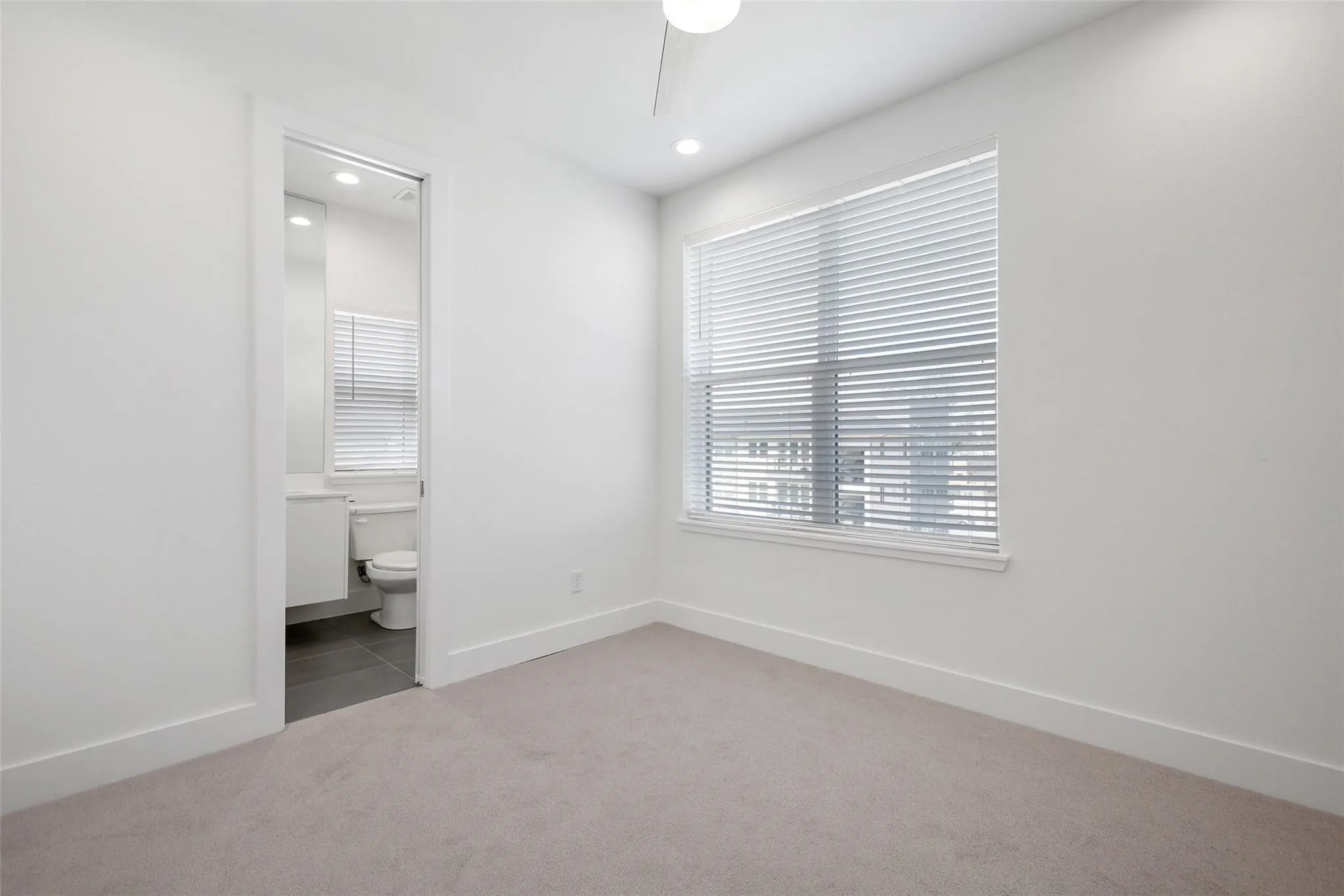 Neutral-toned interior space featuring wall-to-wall carpeting, recessed ceiling lighting, a ceiling fan, a window with horizontal blinds, and access to a bathroom with tile flooring