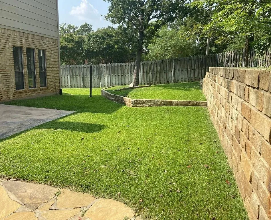 Backyard featuring a tiered stone retaining wall, flagstone patio, manicured lawn, and wood privacy fencing