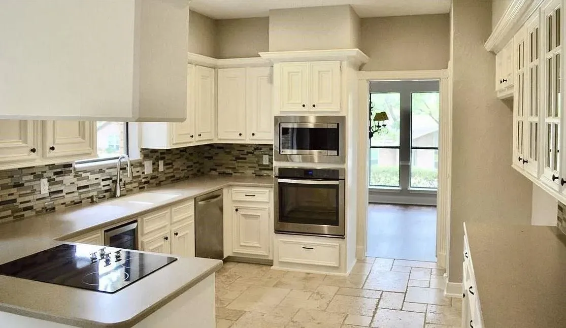 Kitchen featuring white cabinetry, stone-finish tile flooring, stacked-stone backsplash, stainless steel built-in double oven and microwave, and solid-surface countertops