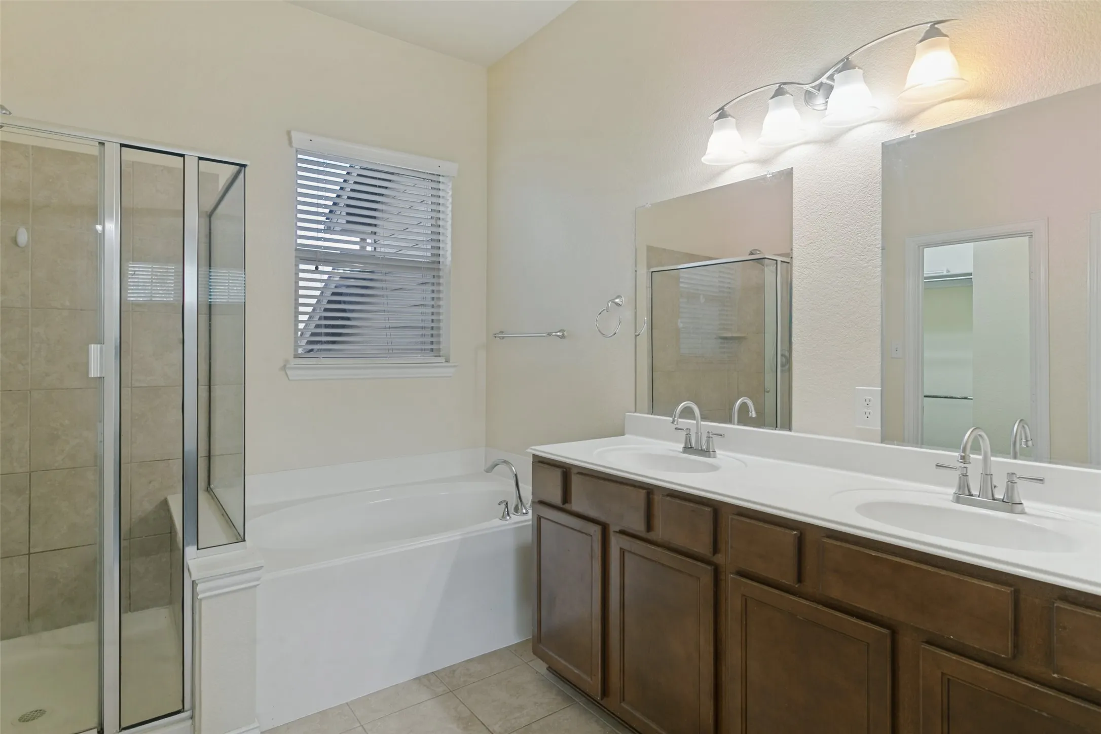 Bathroom featuring a dual-sink vanity with dark-finish cabinetry, a separate soaking tub, a glass-enclosed shower with neutral-tone wall tile, and wall-mounted vanity lighting