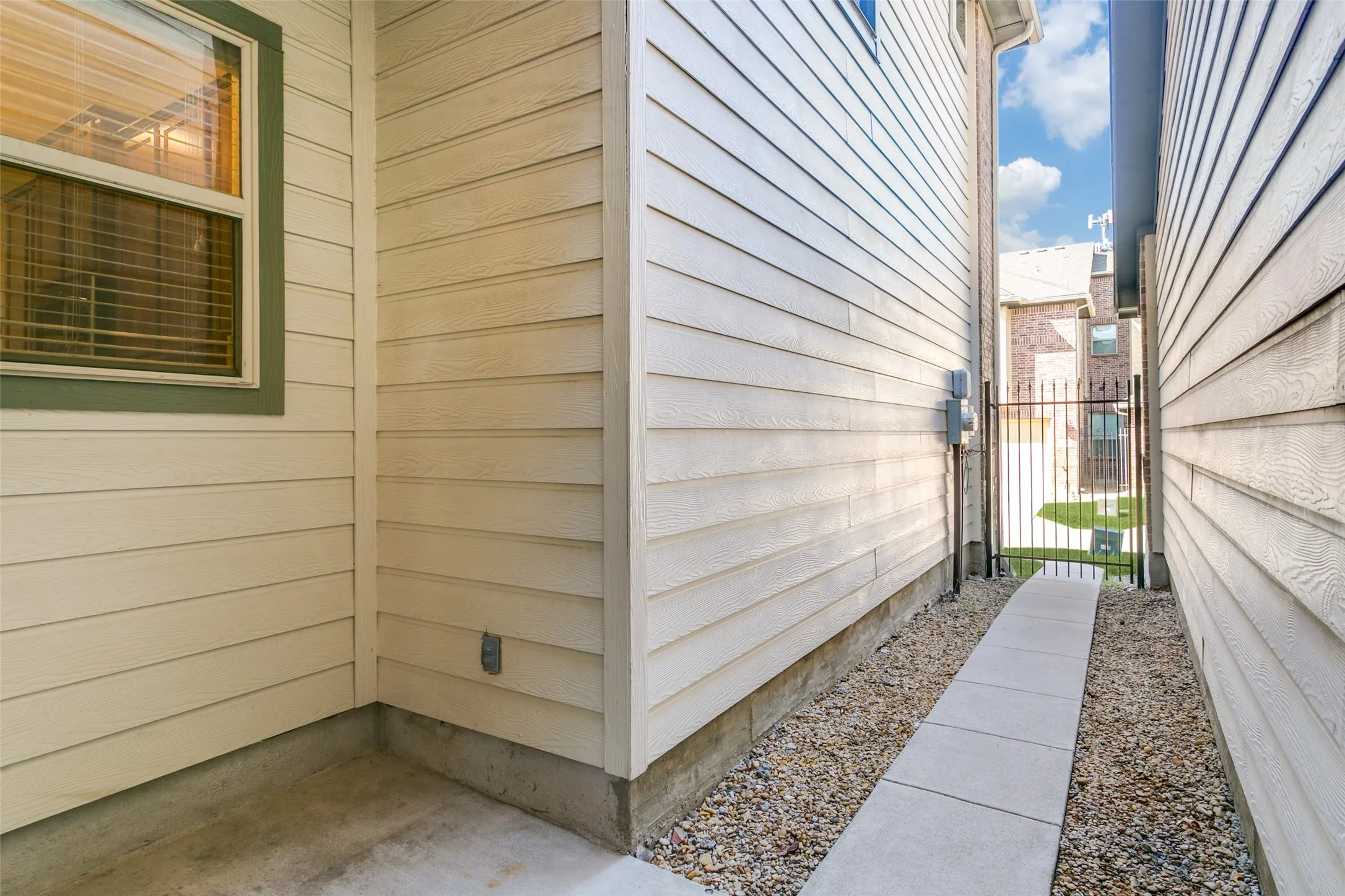 Exterior side passage featuring horizontal lap siding, concrete paving, gravel ground cover, and a metal security gate