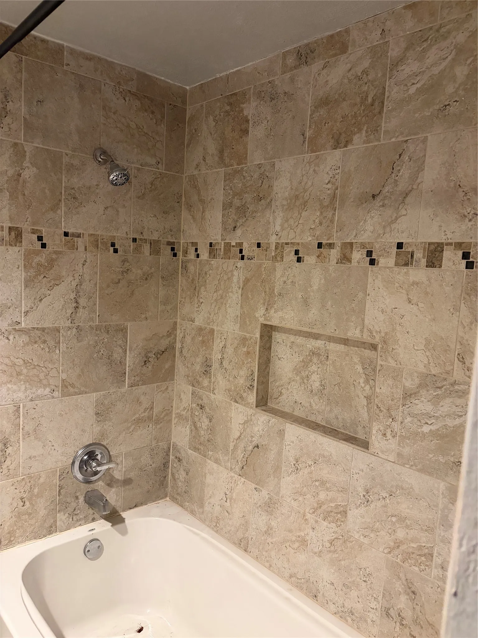 Tub and shower combination featuring floor-to-ceiling stone-look tile, a decorative tile inlay border, a recessed wall niche, and polished chrome fixtures