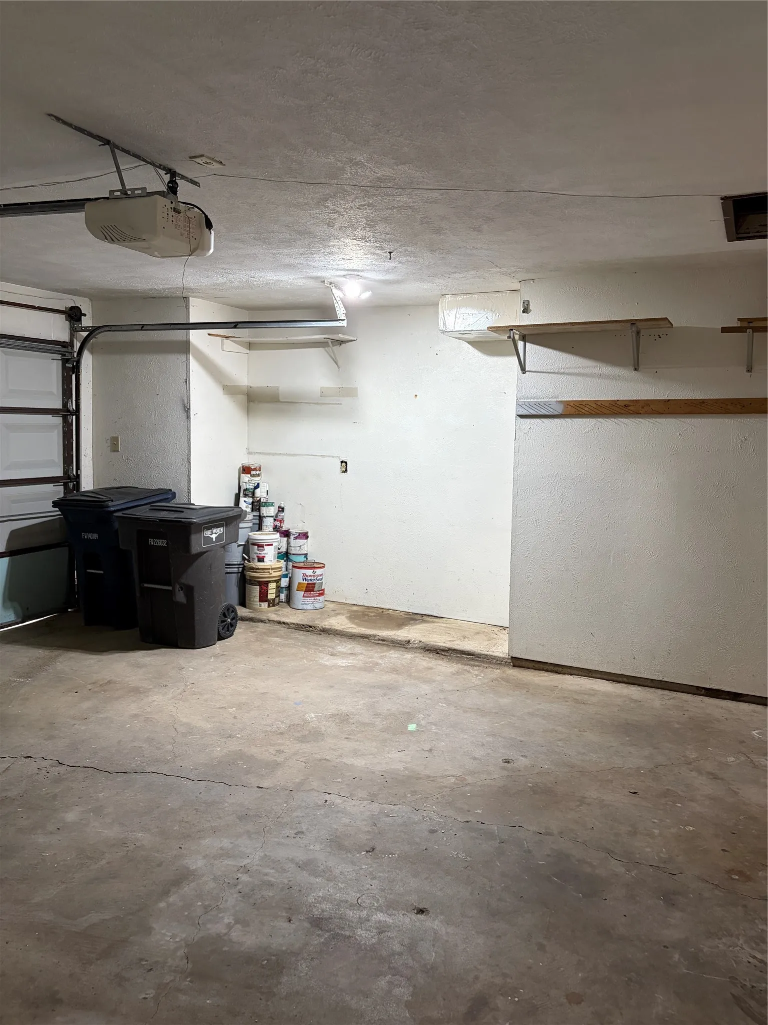 Garage space featuring a concrete floor, motorized overhead garage door opener, and mounted wall shelving