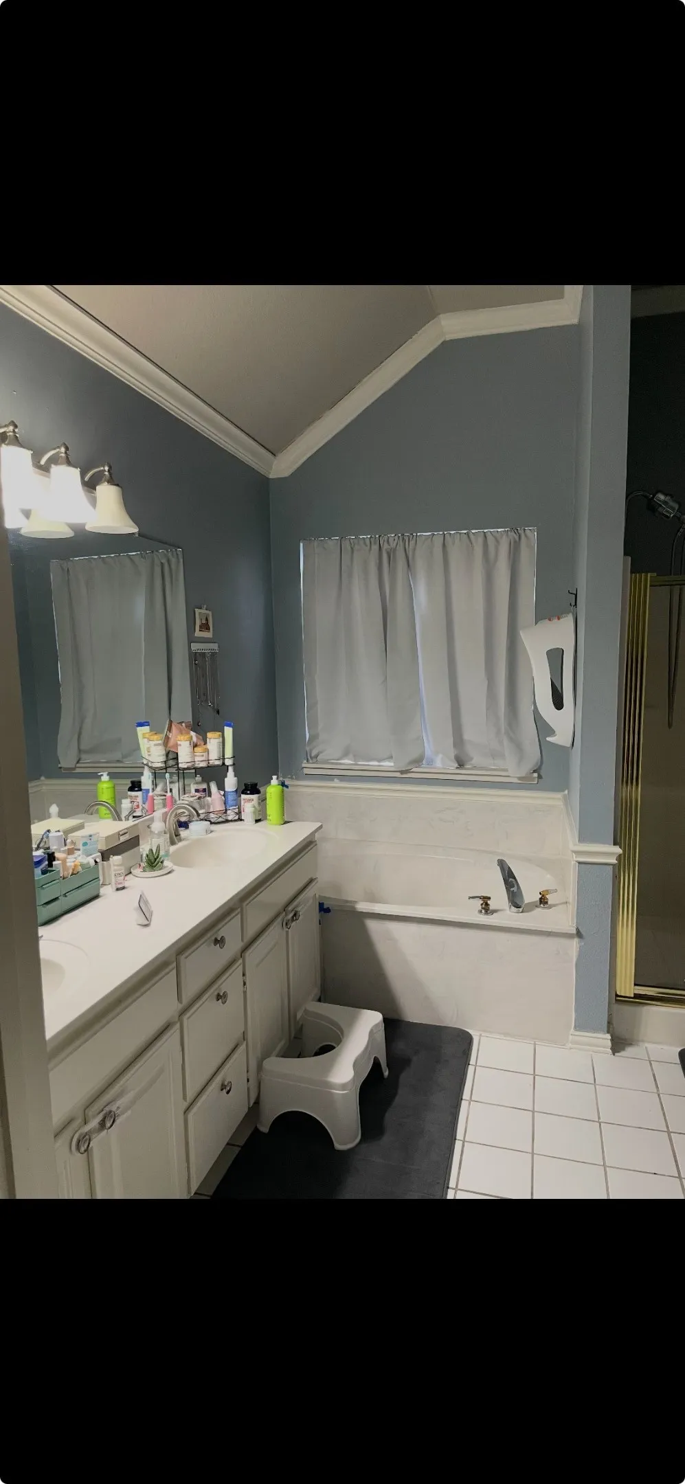 Bathroom featuring a dual-sink vanity with white cabinetry, a drop-in soaking tub with a tiled surround, a separate enclosed shower with gold-tone frame, and vaulted ceilings