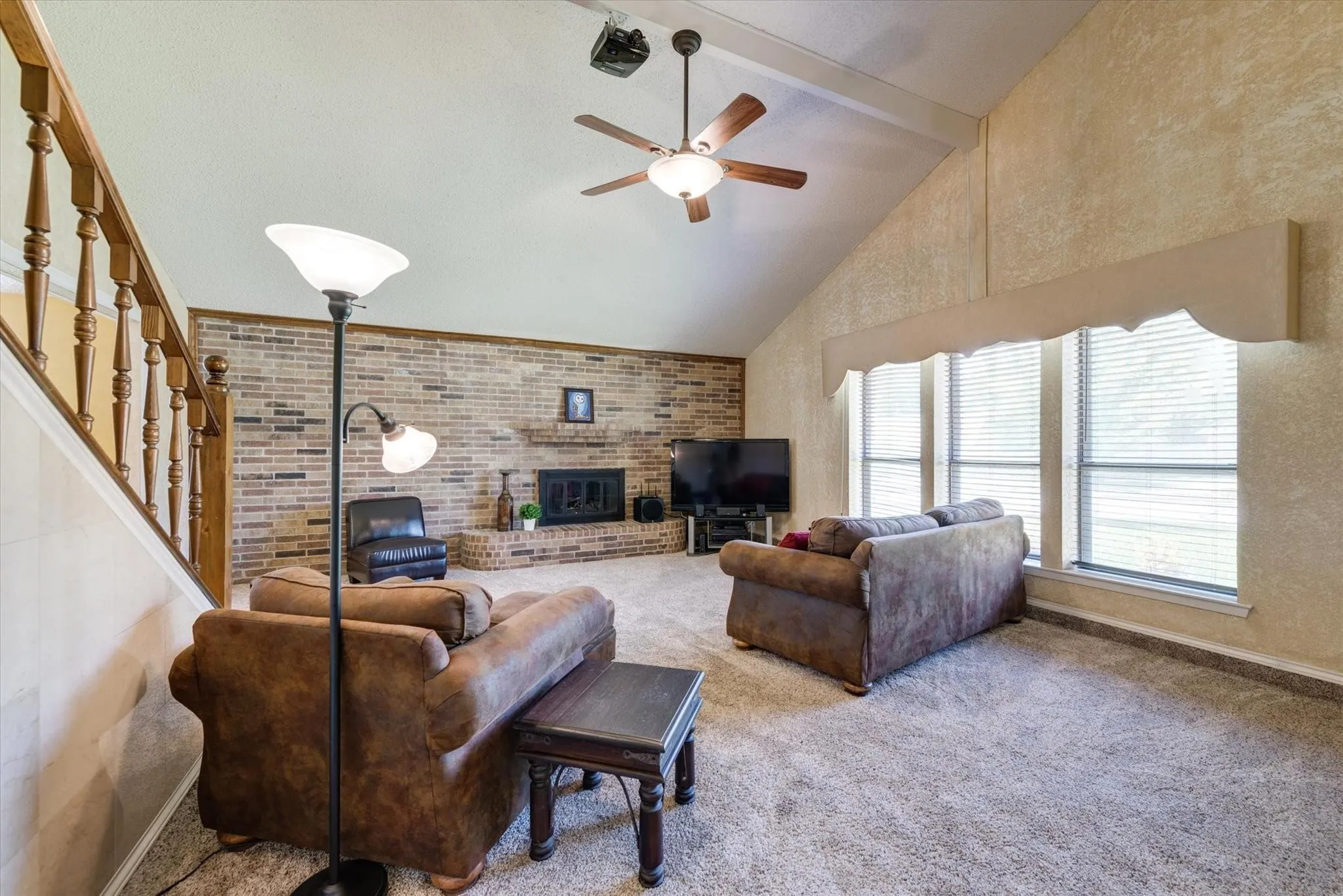Vaulted ceiling featuring a central ceiling fan, exposed brick fireplace with a raised hearth, and wooden staircase railing