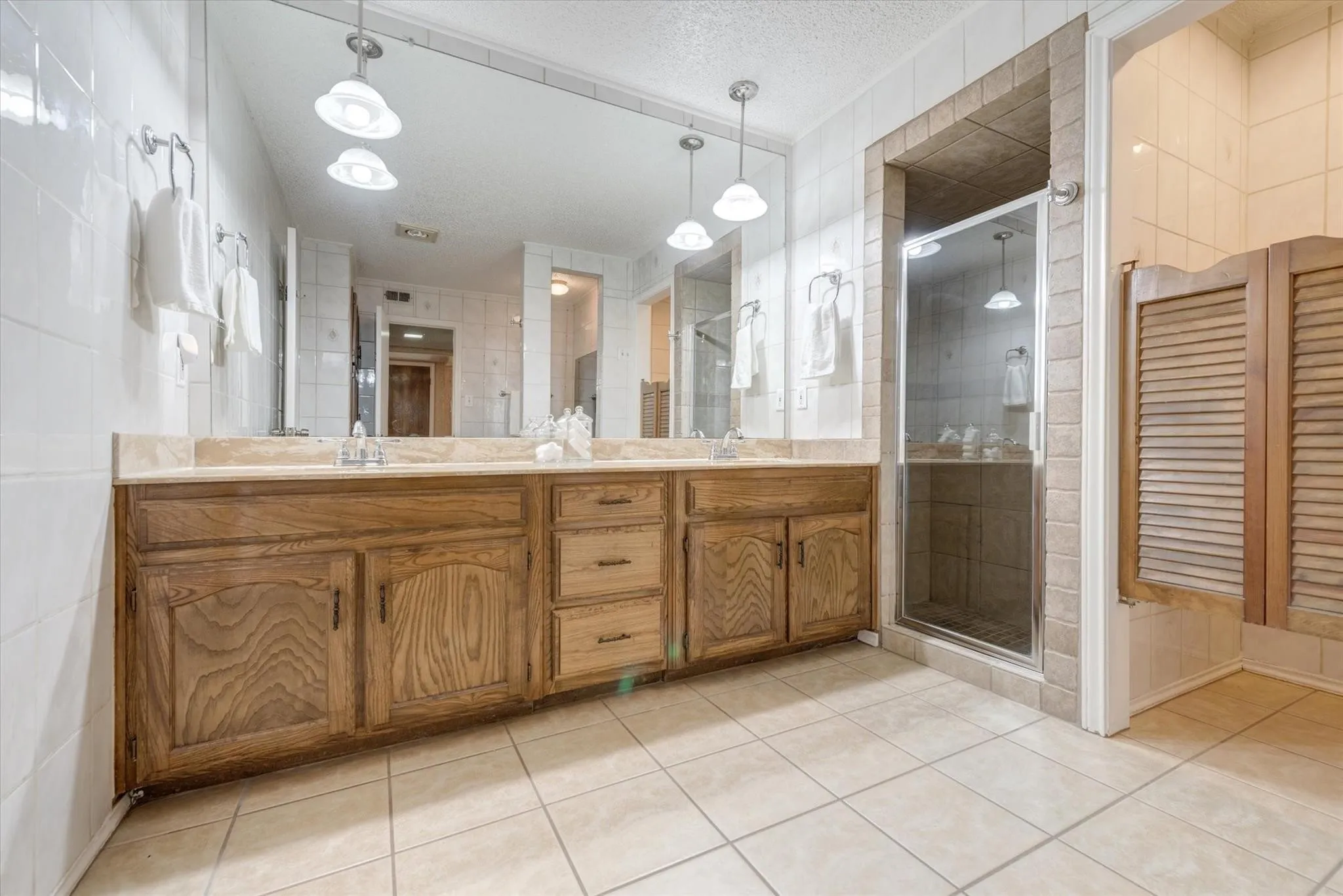 Double-sink wood vanity featuring cabinet storage and drawers, large wall-mounted mirror, pendant lighting, tiled shower enclosure, and ceramic floor tile