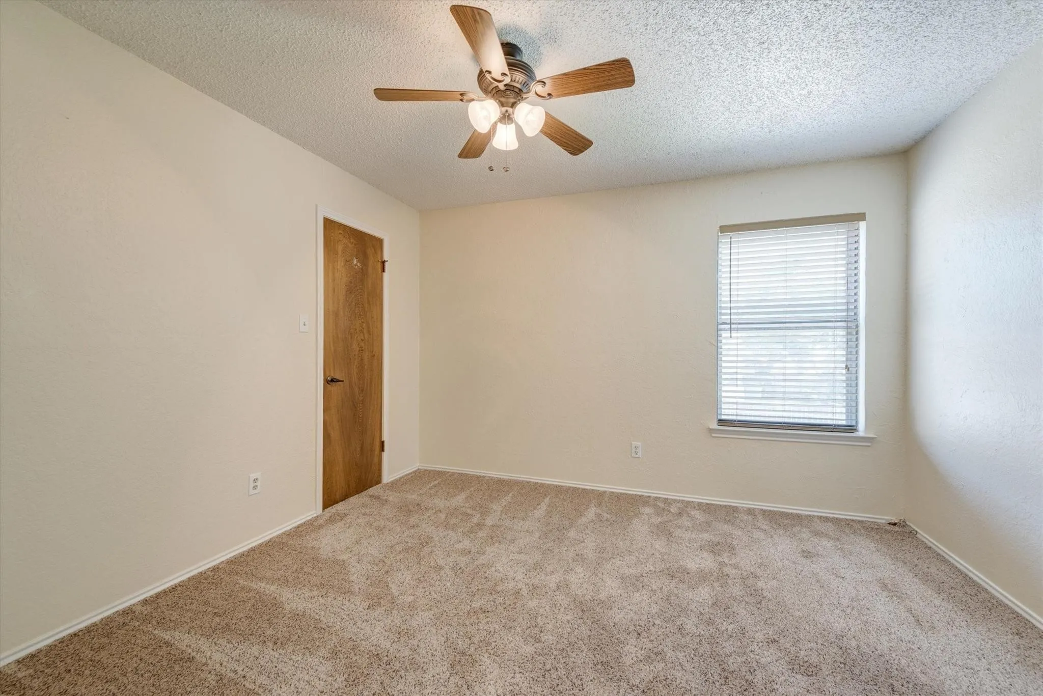 Neutral-toned carpeted room featuring a ceiling fan with integrated lighting, a single-panel wood-grain door, and a window with fitted horizontal blinds