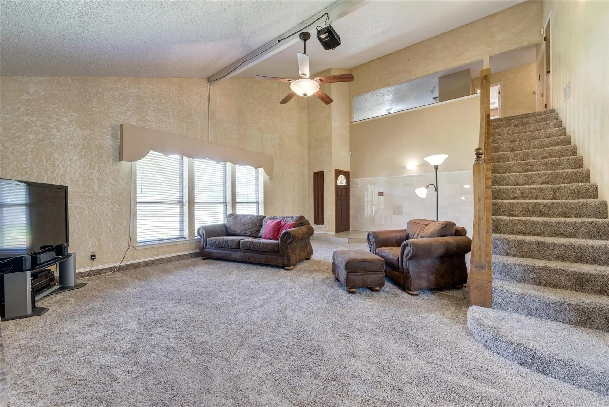 Vaulted ceilings, expansive windows with custom window valances, carpeted staircase featuring a wooden handrail, and ceiling fan with integrated lighting