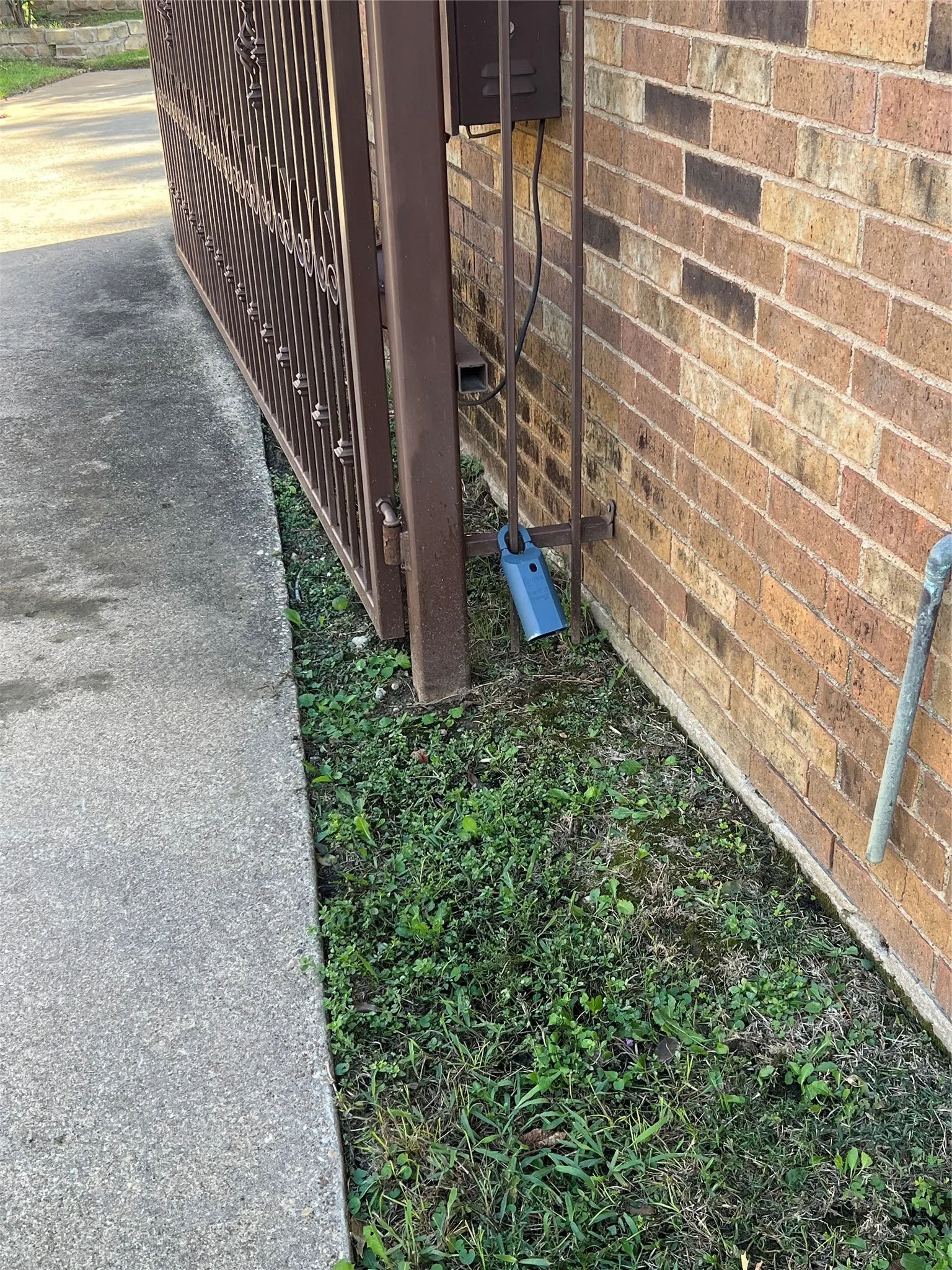 Brown metal security gate with ornate detailing, secured by a heavy-duty padlock, installed against a traditional brick exterior wall