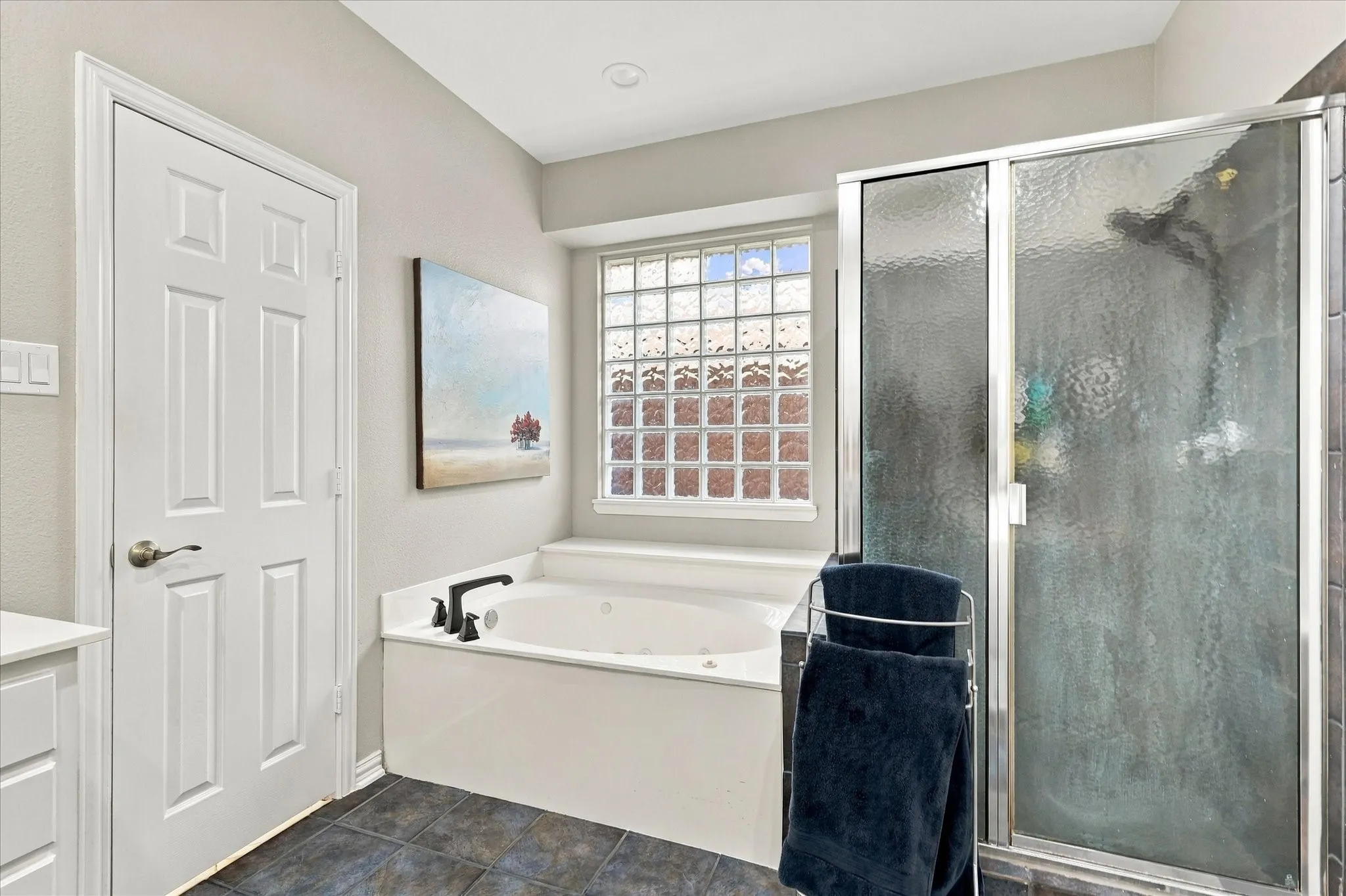 Soaking tub with matte black fixtures, glass block window, glass-enclosed shower, and dark stone-finish floor tile