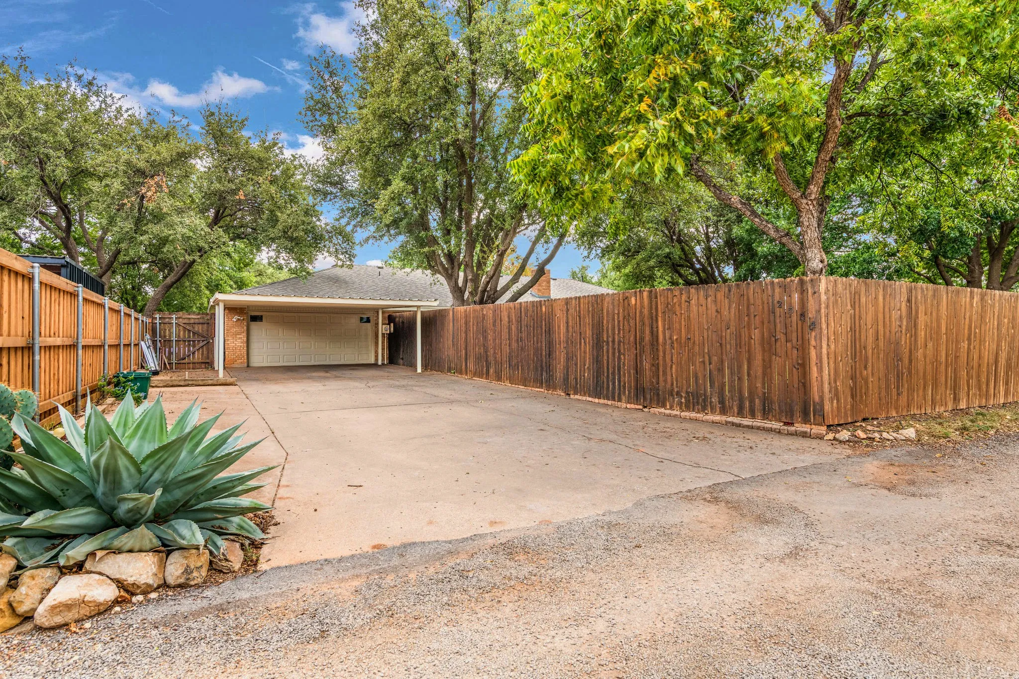 Alley access opens to a broad concrete apron and covered carport leading to the 2 car garage, softened by spiky blue agave, limestone boulders, and a gated side entry in a stout cedar-toned fence.