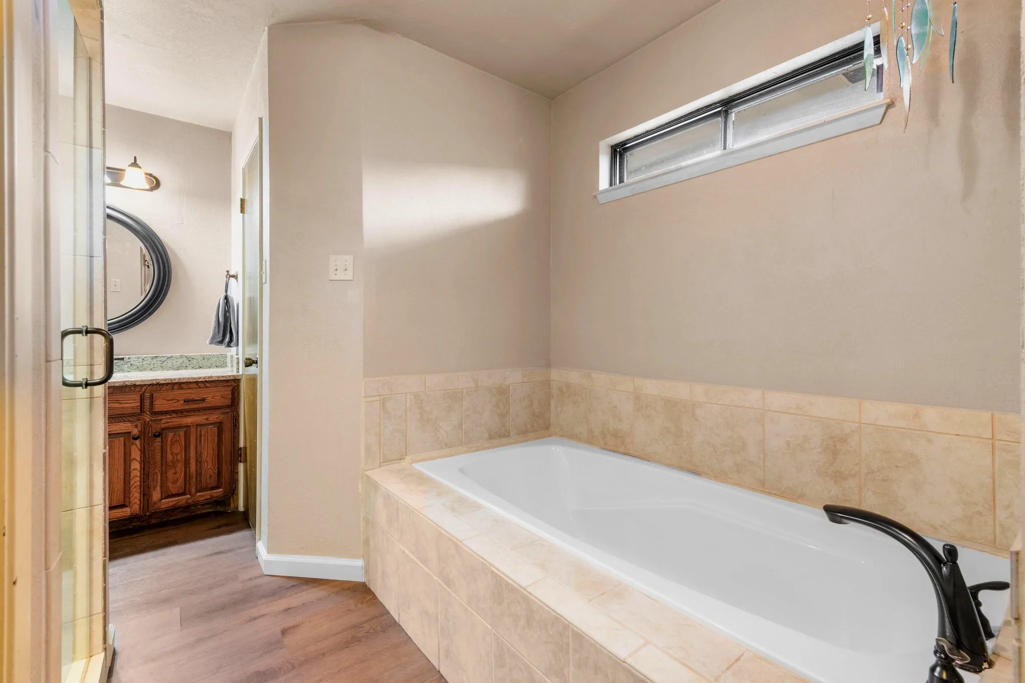 A deep soaking tub wrapped in sand-colored tile sits beneath a slim transom window, where a glass wind chime catches the daylight