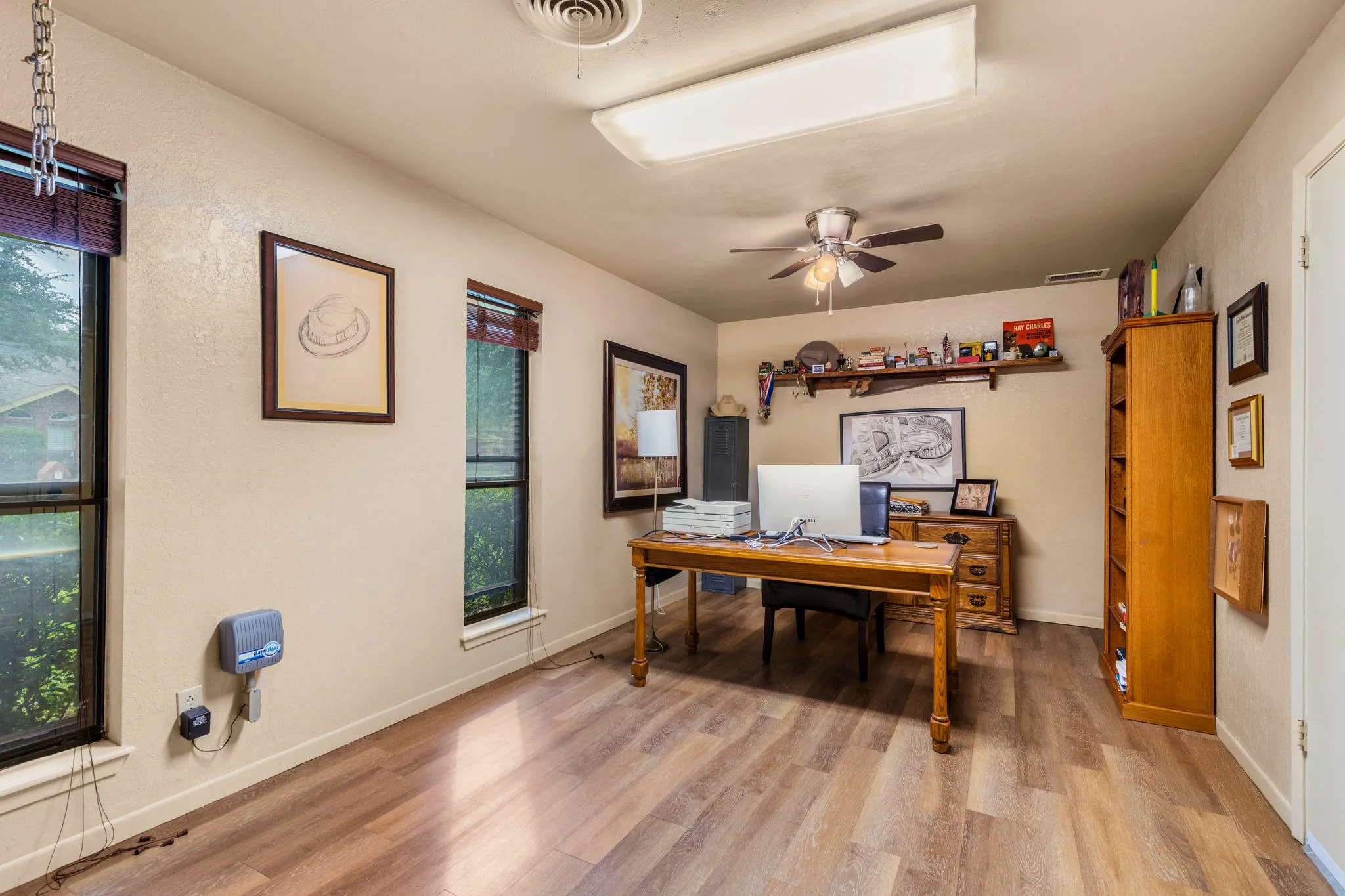 Natural light floods this quiet study through tall windows that frame a garden of greenery, while soft neutral walls and a rich wood-look surface underfoot create a calm place to work, read, or dream