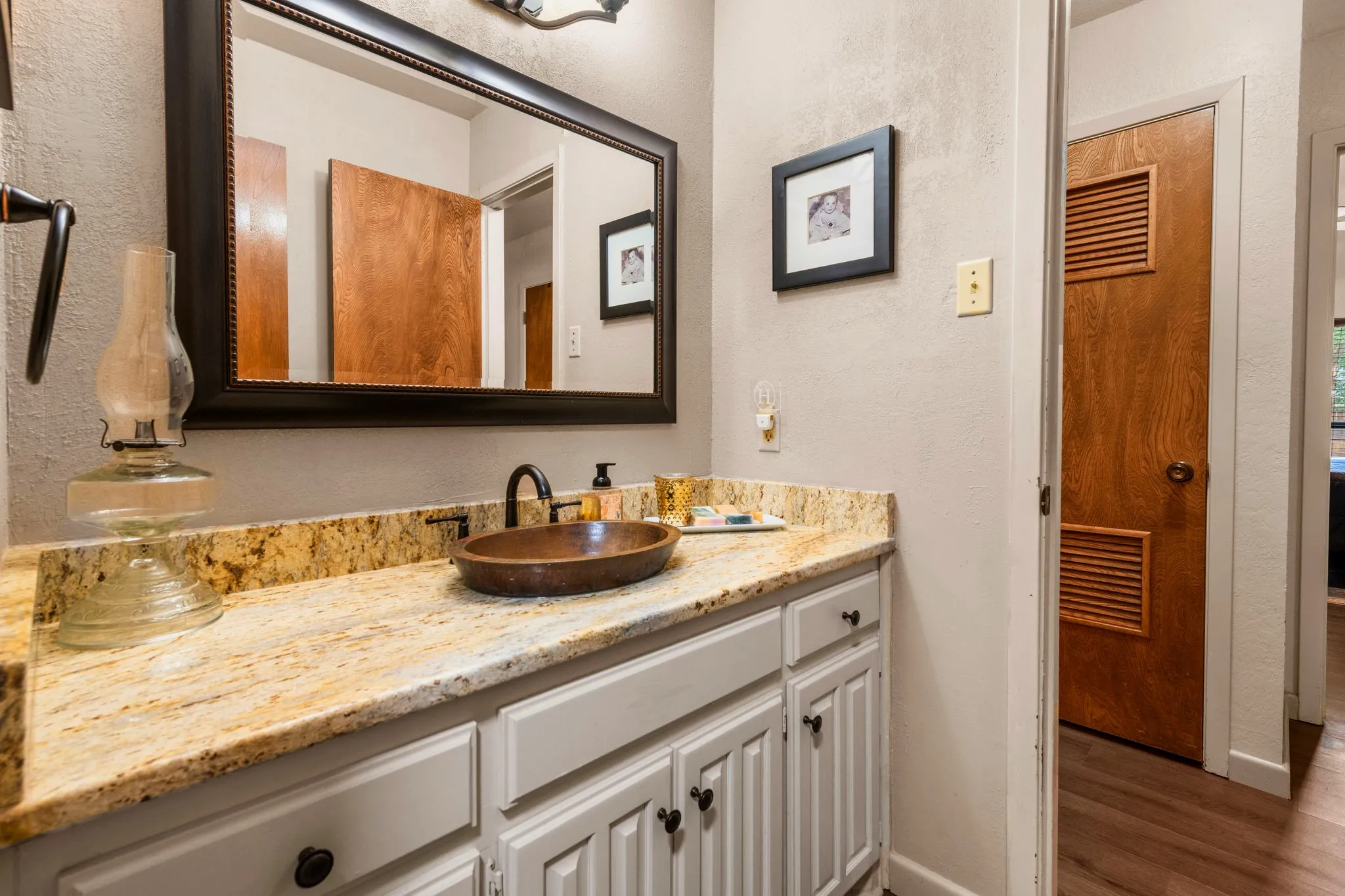 A generous mirror in a dark, sculpted frame edged with delicate beaded trim reflects the room above ivory raised-panel cabinetry, finished with dark knobs and topped by a polished granite counter swirled in amber, caramel, and umber.