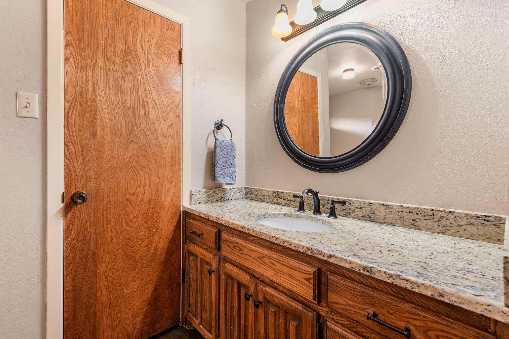 An oversized round mirror in a dusky black frame crowns a speckled cream-and-charcoal countertop, an undermount basin, and richly stained oak cabinetry