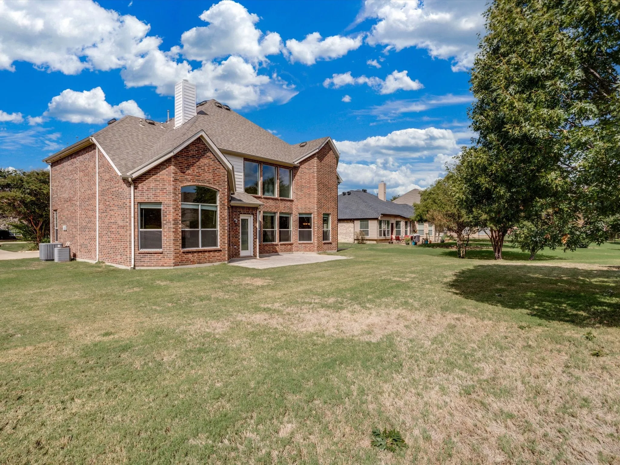 Two-story brick exterior featuring a multi-gabled roofline, expansive lawn, concrete patio, and large glass-paneled windows